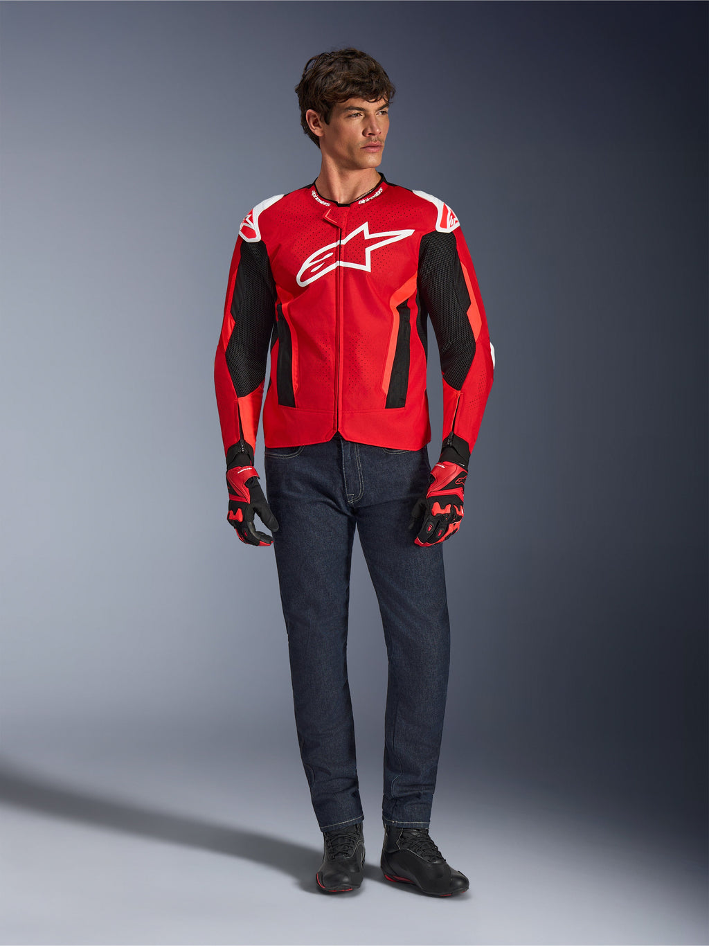 Kurtka Alpinestars T-GP Air, Bright Red Red Fluo Black, męski model w pełnym stroju motocyklowym z ciemnoniebieskimi dżinsami i pasującymi rękawicami, sportowa kurtka tekstylna z perforowanymi panelami z siatki i białym logo Astars, zdjęcie studyjne.