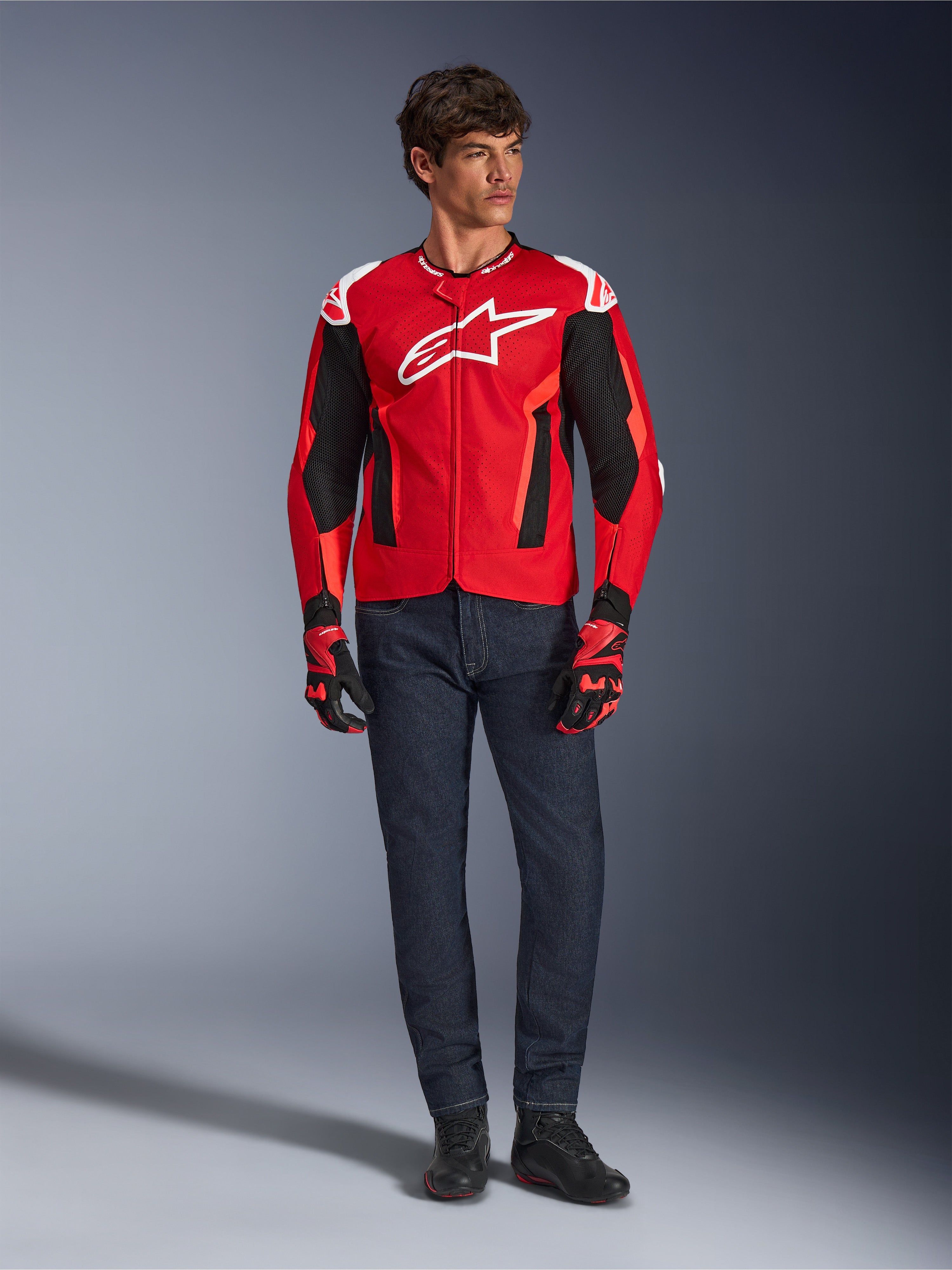 Kurtka Alpinestars T-GP Air, Bright Red Red Fluo Black, męski model w pełnym stroju motocyklowym z ciemnoniebieskimi dżinsami i pasującymi rękawicami, sportowa kurtka tekstylna z perforowanymi panelami z siatki i białym logo Astars, zdjęcie studyjne.