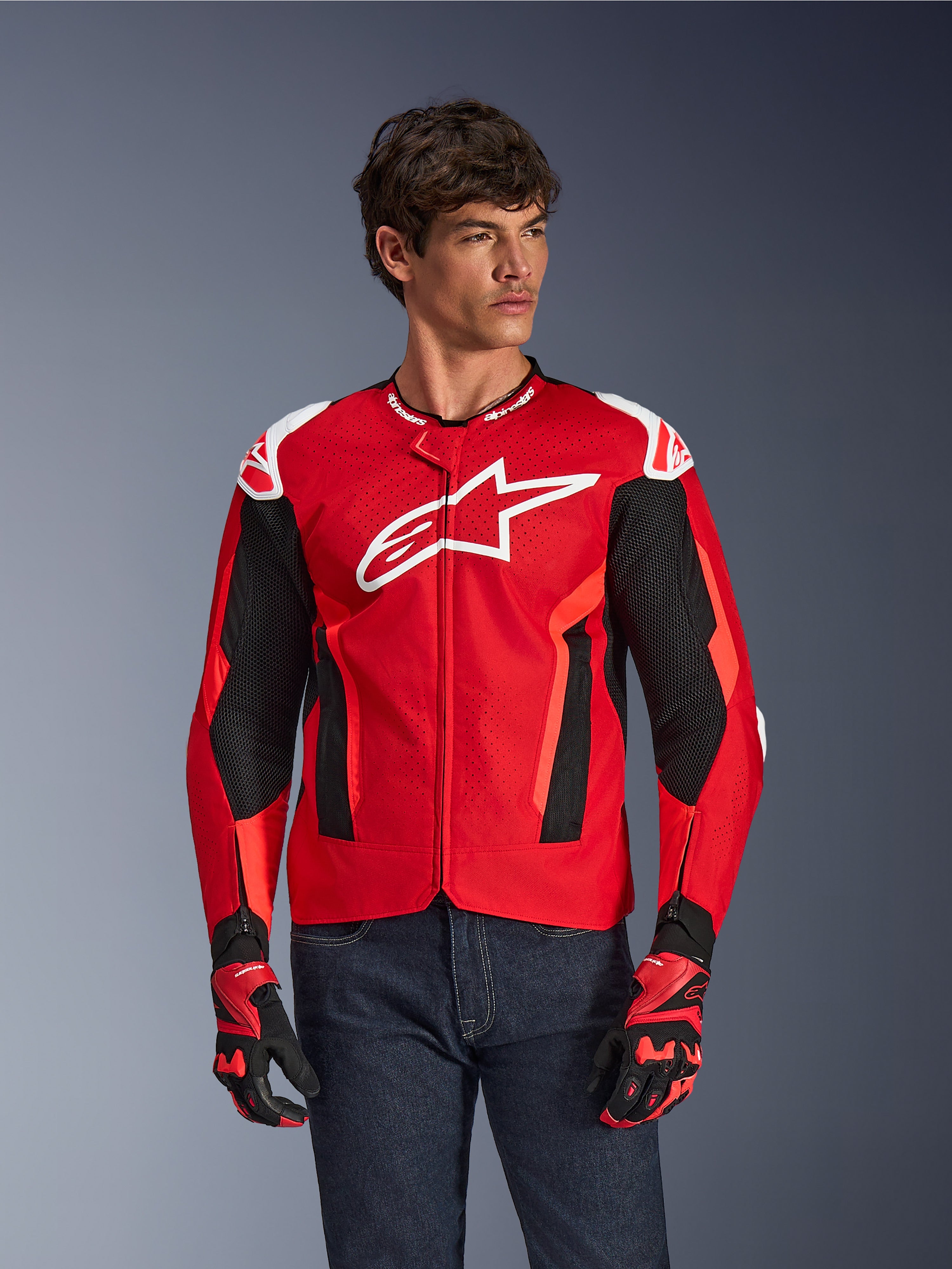 Alpinestars T-GP Air Jacket, kurtka motocyklowa, jaskrawoczerwona i czarna z białym logo, z perforowanymi panelami wentylacyjnymi z siatki, noszona z dopasowanymi rękawicami i ciemnymi dżinsami, sportowy design do jazdy w ciepłe dni