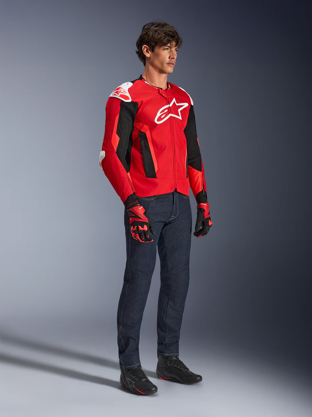Kurtka Alpinestars T-GP Air, sportowa kurtka motocyklowa, jaskrawoczerwona i czarna z białymi akcentami, prezentowana przez modela w ciemnoniebieskich dżinsach i czerwonych rękawicach wyścigowych, z panelami z siatki o wysokim przepływie powietrza i białymi sliderami na ramionach, aerodynamiczny sportowy krój.