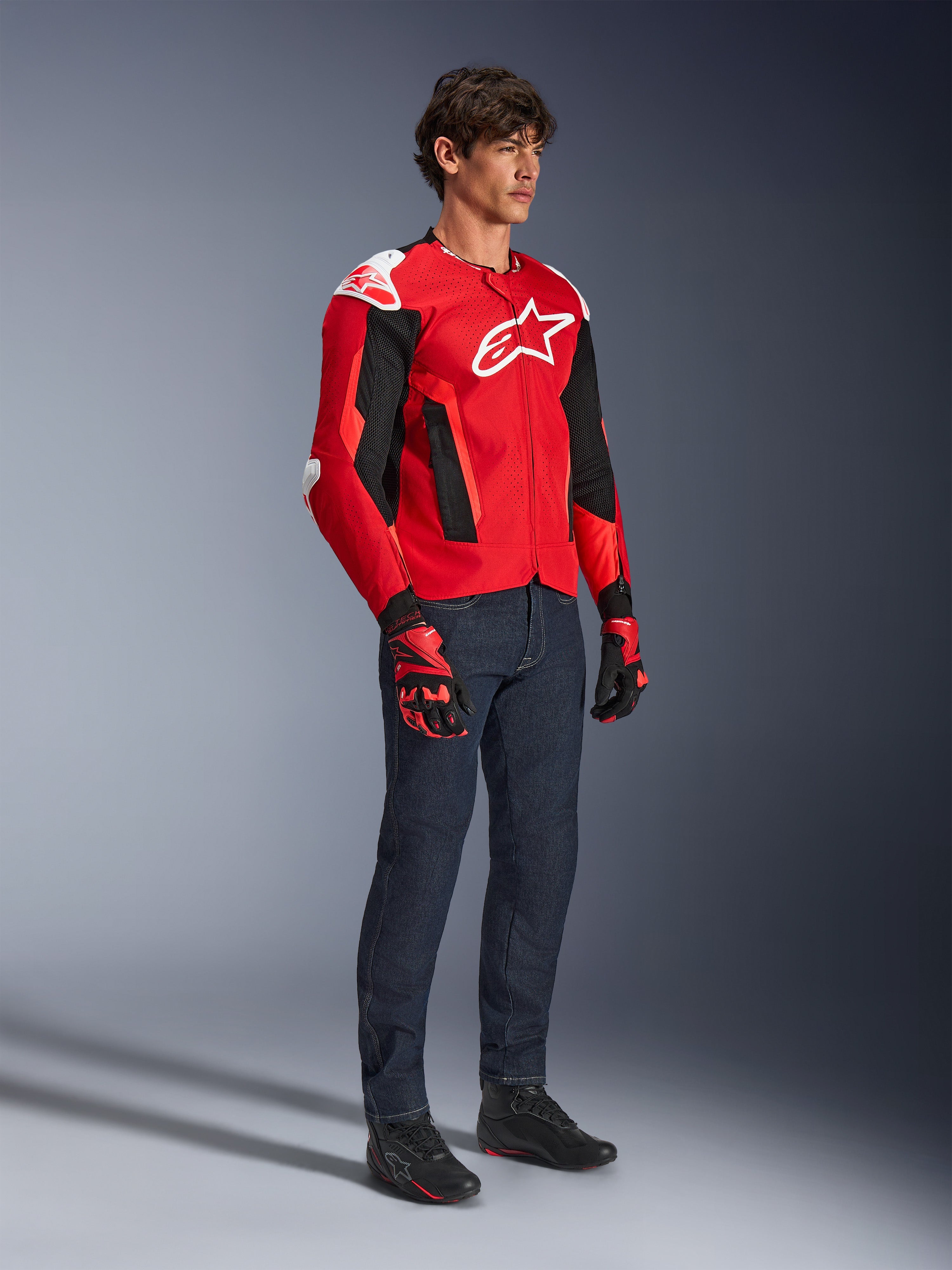 Kurtka Alpinestars T-GP Air, sportowa kurtka motocyklowa, jaskrawoczerwona i czarna z białymi akcentami, prezentowana przez modela w ciemnoniebieskich dżinsach i czerwonych rękawicach wyścigowych, z panelami z siatki o wysokim przepływie powietrza i białymi sliderami na ramionach, aerodynamiczny sportowy krój.