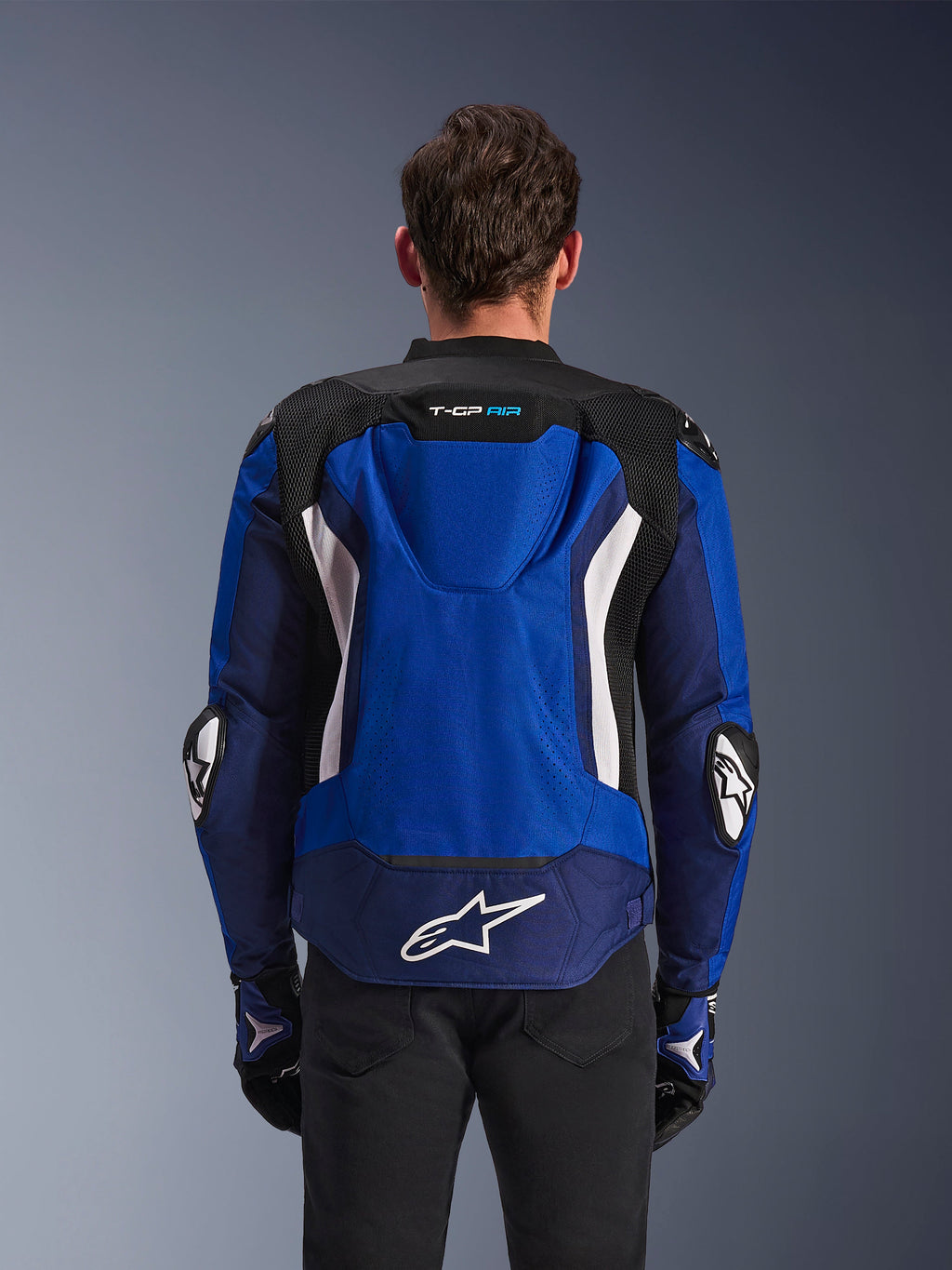 Alpinestars T-GP Air Jacket, sportowa tekstylna kurtka motocyklowa, jasnoniebieska, granatowa i biała, widok z tyłu prezentujący zintegrowany garb aerodynamiczny i duże białe logo w dolnej części pleców, noszona z dopasowanymi niebiesko-czarnymi rękawicami motocyklowymi