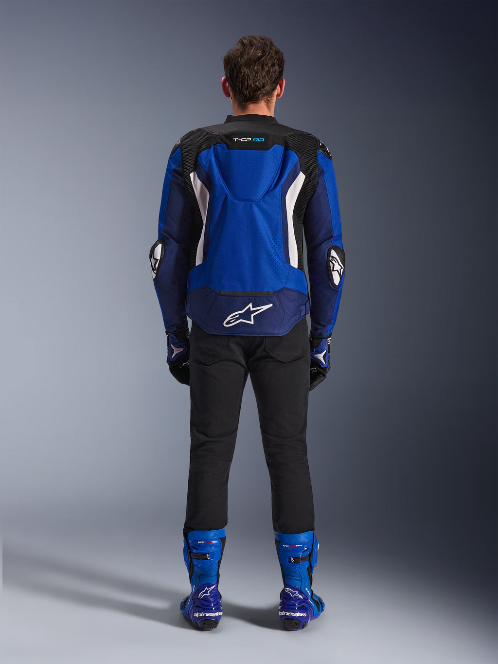 Alpinestars T-GP Air Jacket, jasnoniebieska, granatowa i biała, widok z tyłu prezentujący garb aerodynamiczny i siatkowe panele wentylacyjne, białe logo Alpinestars w dolnej części pleców, zestawiona z czarnymi spodniami i niebieskimi butami wyścigowymi
