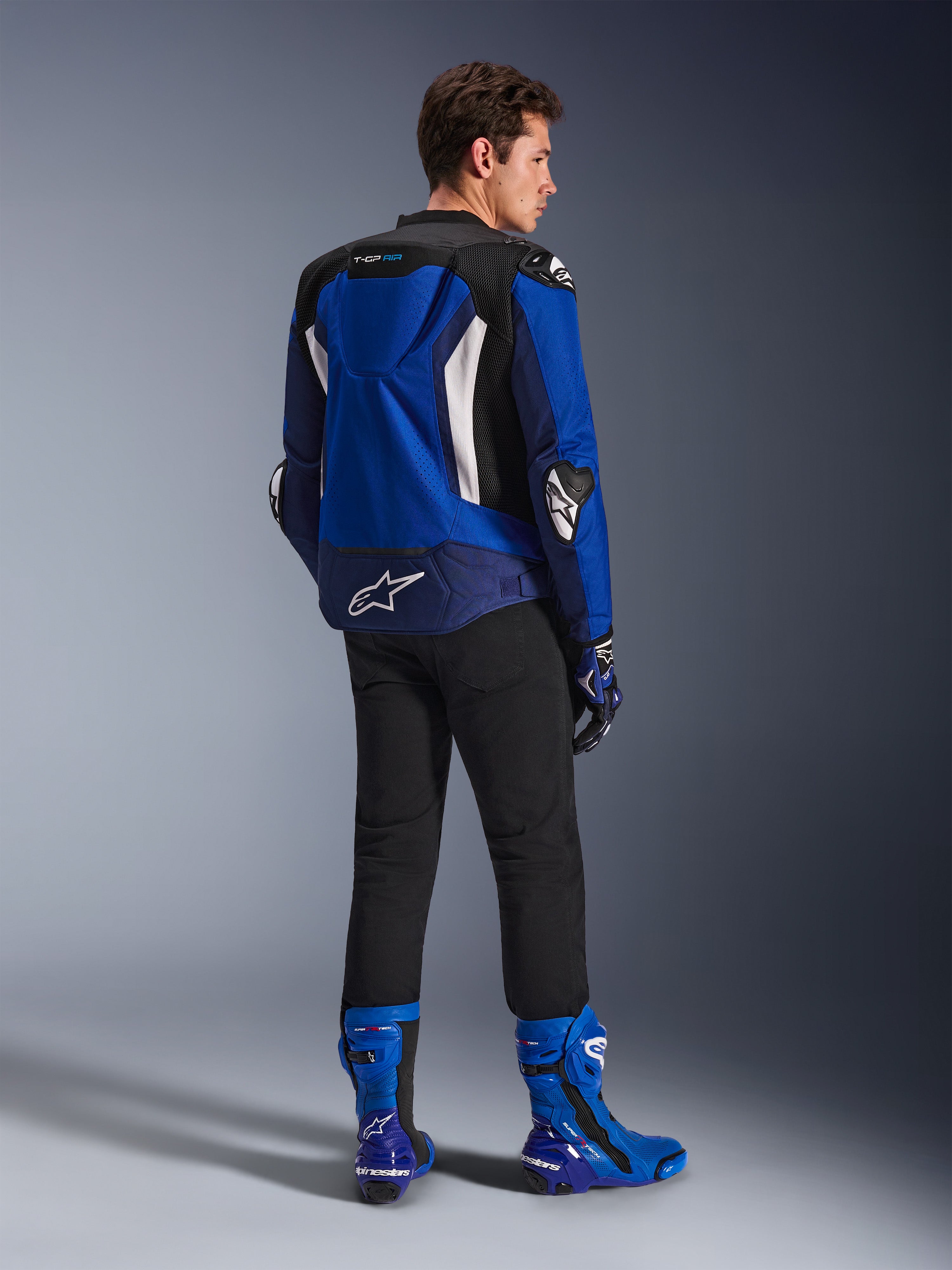 Kurtka Alpinestars T-GP Air, sportowa kurtka tekstylna, jasnoniebieska, granatowa i biała, widok z tyłu na modelu w czarnych spodniach i pasujących niebiesko-białych butach, białe logo Alpinestars na dole pleców, panele wentylacyjne z siatki i garb aerodynamiczny