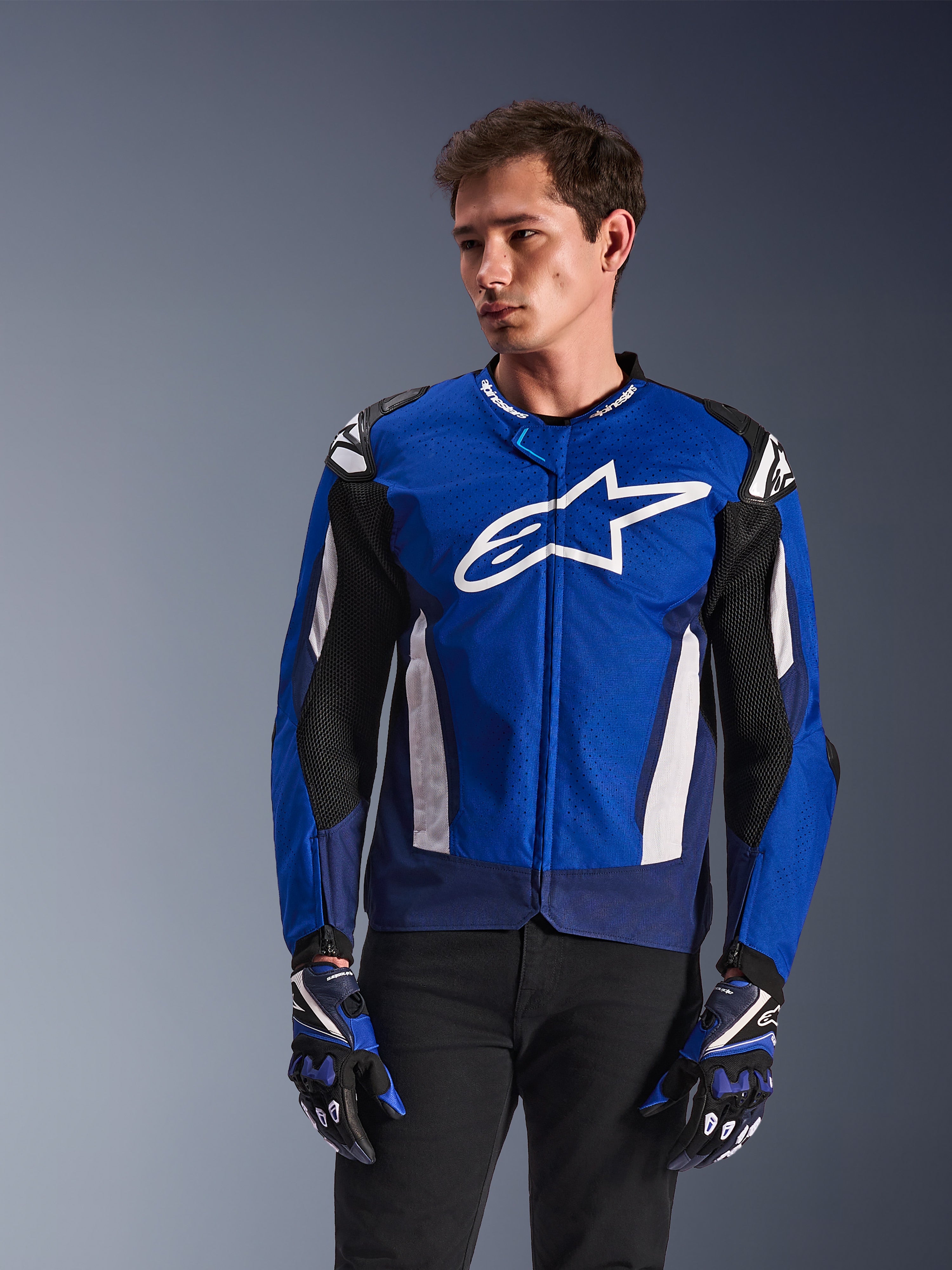Alpinestars T-GP Air Jacket, sportowa tekstylna kurtka motocyklowa, jasnoniebieska i granatowa z białymi akcentami, z dużym logo gwiazdy na piersi, siateczkowe panele wentylacyjne, noszona przez modela z pasującymi rękawicami i czarnymi spodniami