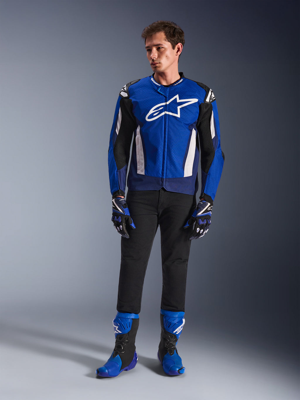 Męski model ubrany w kurtkę Alpinestars T-GP Air w kolorze Bright Blue Navy White, czarne spodnie, dopasowane niebiesko-granatowe rękawice wyścigowe oraz niebiesko-czarne buty motocyklowe, zdjęcie studyjne prezentujące kompletny sportowy strój motocyklowy.