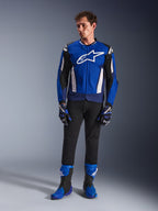 Męski model ubrany w kurtkę Alpinestars T-GP Air w kolorze Bright Blue Navy White, czarne spodnie, dopasowane niebiesko-granatowe rękawice wyścigowe oraz niebiesko-czarne buty motocyklowe, zdjęcie studyjne prezentujące kompletny sportowy strój motocyklowy.