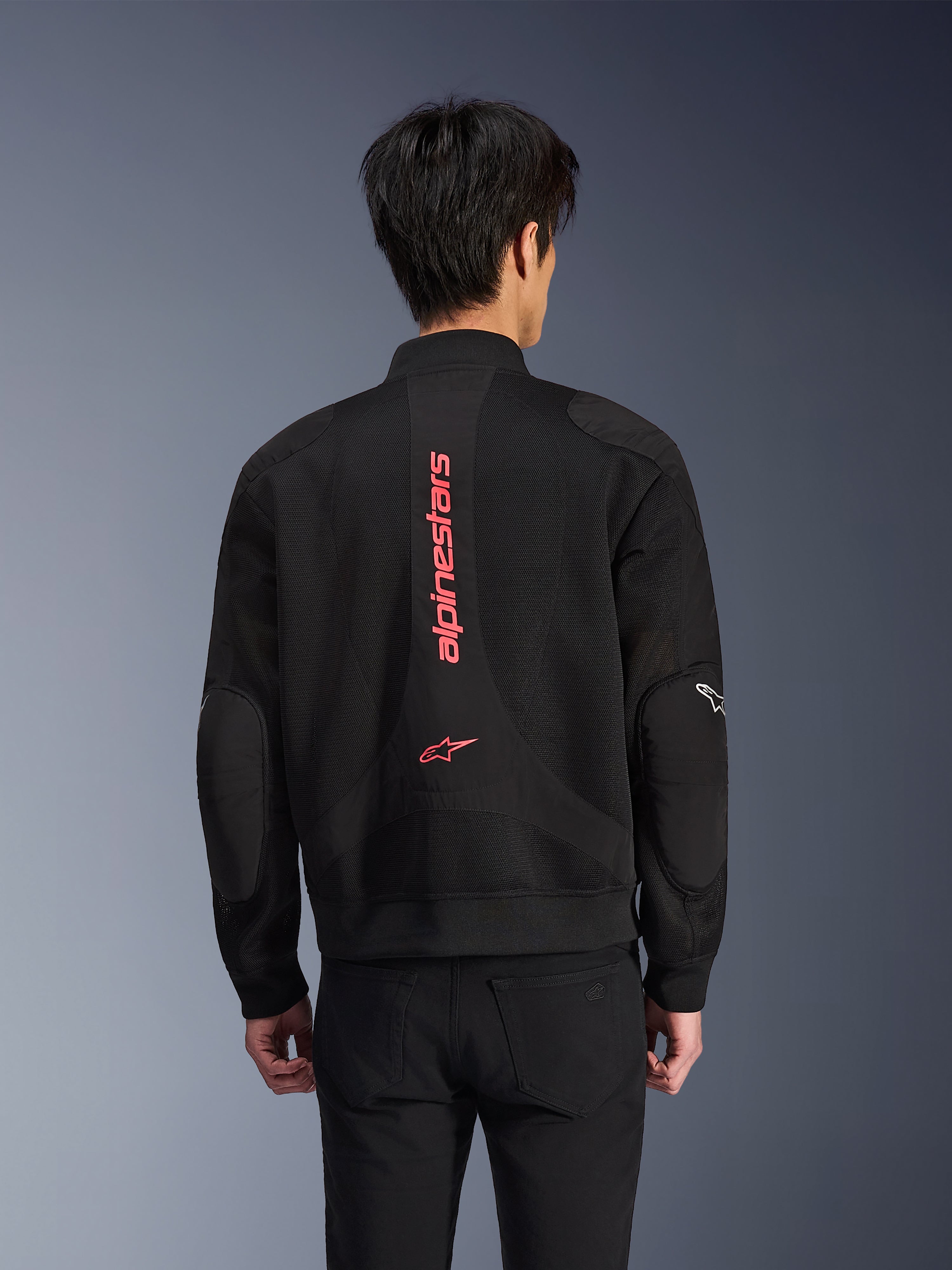 Widok od tyłu motocyklisty w kurtce Alpinestars Flight Air w kolorze Black Bright Red, miejska kurtka motocyklowa w stylu bomber z konstrukcją z siatki 3D, z dużym pionowym czerwonym napisem Alpinestars i logo gwiazdy na plecach, odzież techniczna inspirowana stylem ulicznym
