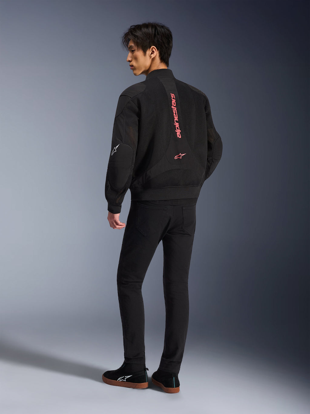 Męski model ubrany w kurtkę Alpinestars Flight Air w kolorze Black Bright Red, widok z tyłu ukazujący pionowe czerwone logo na plecach oraz konstrukcję z siatki 3D, w połączeniu z czarnymi spodniami i sneakersami, miejski styl motocyklowy