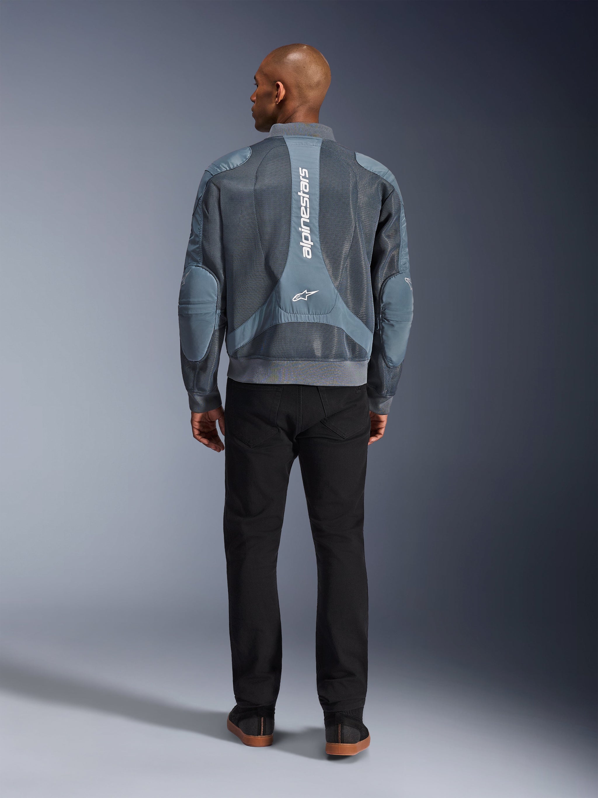 Kurtka Alpinestars Flight Air w kolorze Storm Gray, widok z tyłu na modela w miejskiej kurtce motocyklowej z panelami z siatki 3D i pionowym logo Alpinestars, w zestawieniu z czarnymi spodniami tworzącymi nowoczesny, miejski styl.