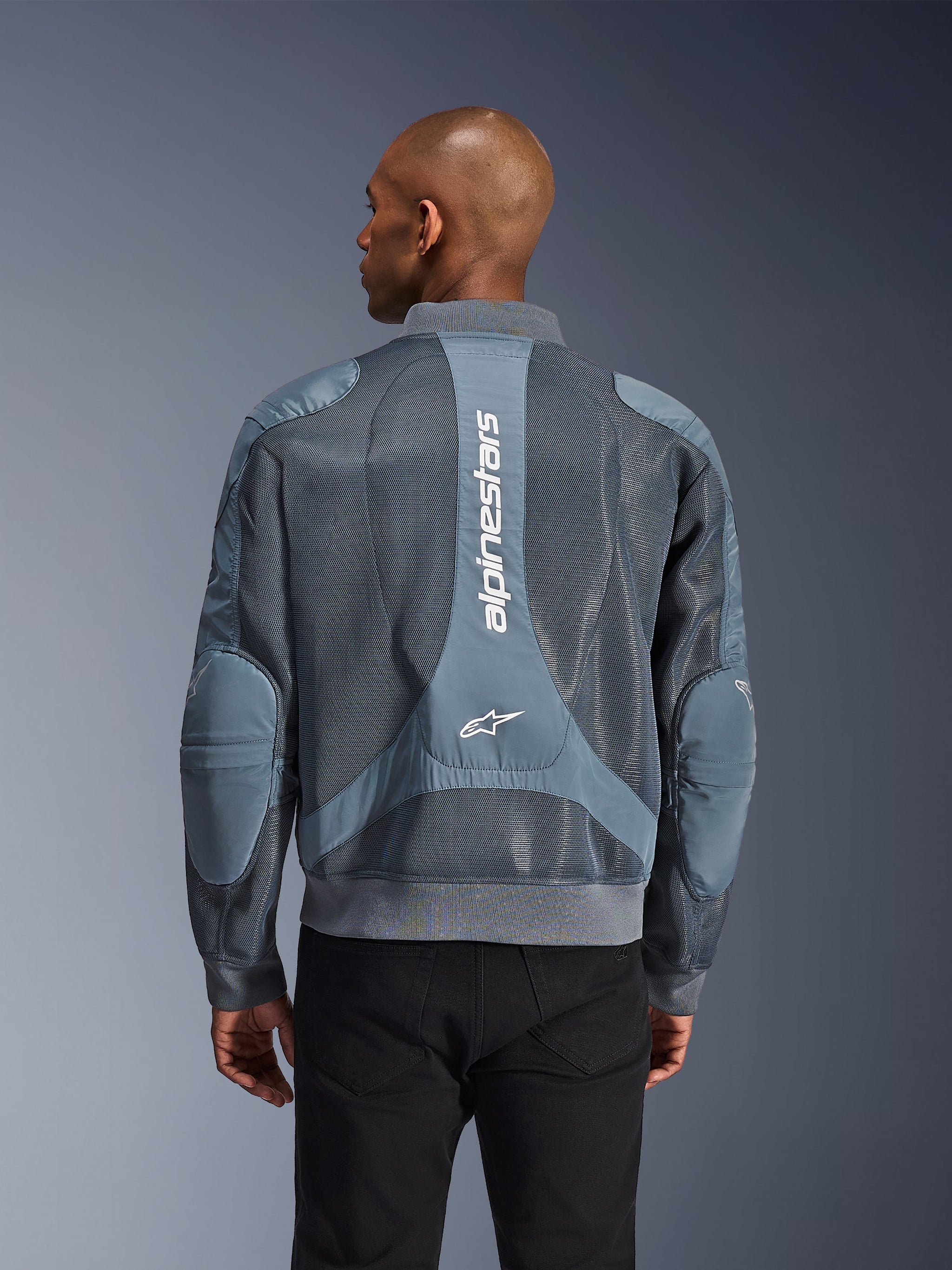 Alpinestars Flight Air Jacket w kolorze Storm Gray, widok z tyłu na modelu, miejska kurtka motocyklowa z rozszerzonymi panelami z siatki 3D, pionowym logo Alpinestars na plecach oraz ściągaczami u dołu i przy kołnierzu w stylu bomberki.