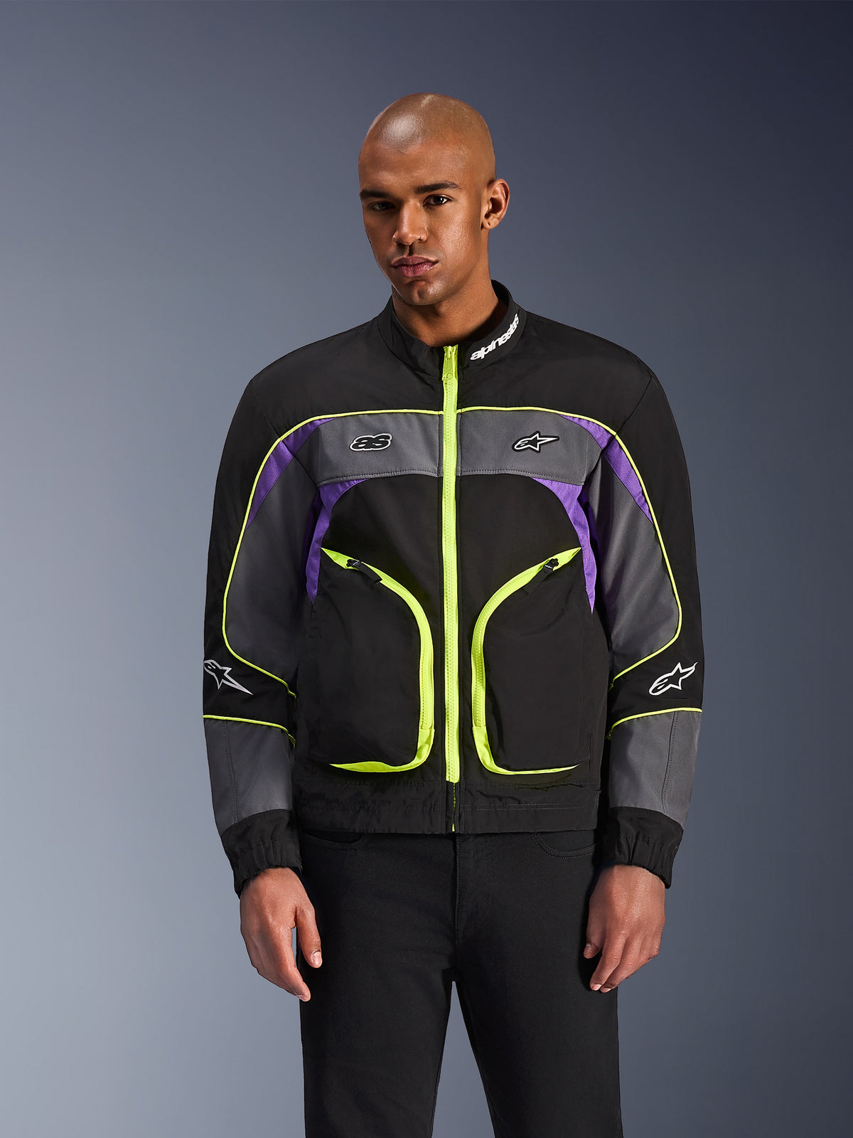 Alpinestars Unite Jacket, miejska kurtka motocyklowa, czarna (Black Asphalt) i fioletowa z neonowożółtymi akcentami, duże przednie kieszenie z neonowymi zamkami, wysoki kołnierz z białym logo, prezentowana przez modela, nowoczesna odzież ochronna inspirowana stylem streetwear