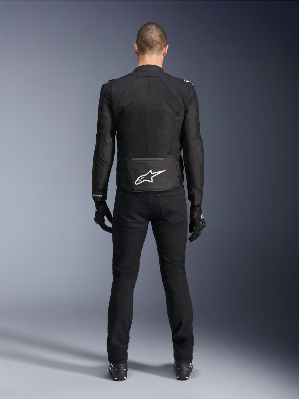 Kurtka Alpinestars Viper Air V4, czarna tekstylna kurtka motocyklowa, widok z tyłu z białym logo gwiazdy Alpinestars w dolnej części pleców, noszona z czarnymi spodniami i rękawicami motocyklowymi, letnia odzież do jazdy szosowej i sportowej