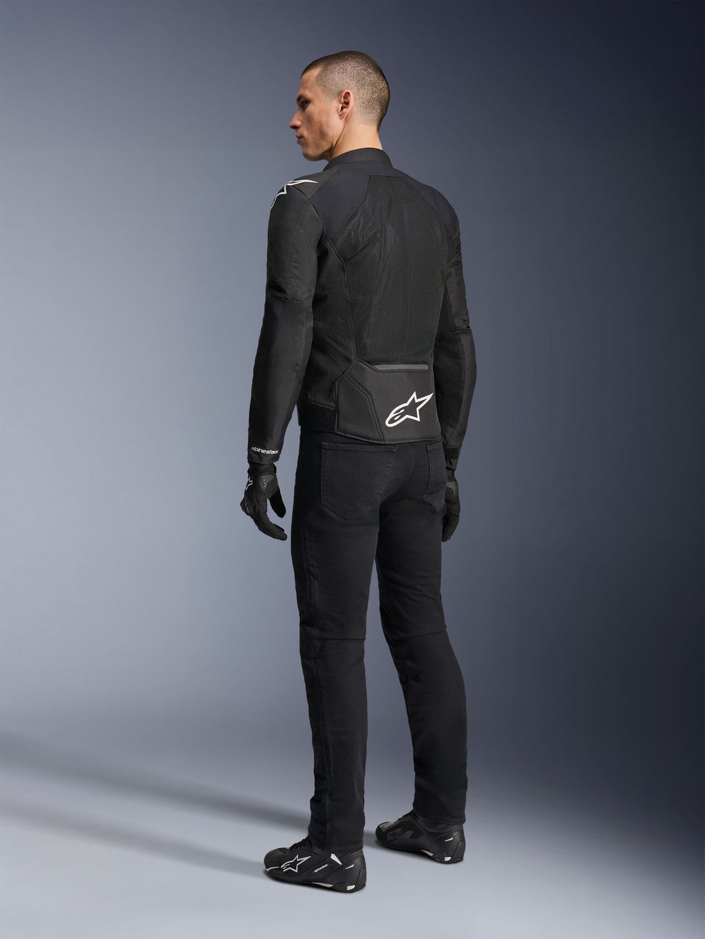 Kurtka Alpinestars Viper Air V4, czarna tekstylna kurtka motocyklowa, widok z tyłu na męskim modelu, duże panele z siatki technologicznej dla optymalnego przepływu powietrza, białe logo gwiazdy na dole pleców, noszona z czarnymi spodniami i rękawicami motocyklowymi