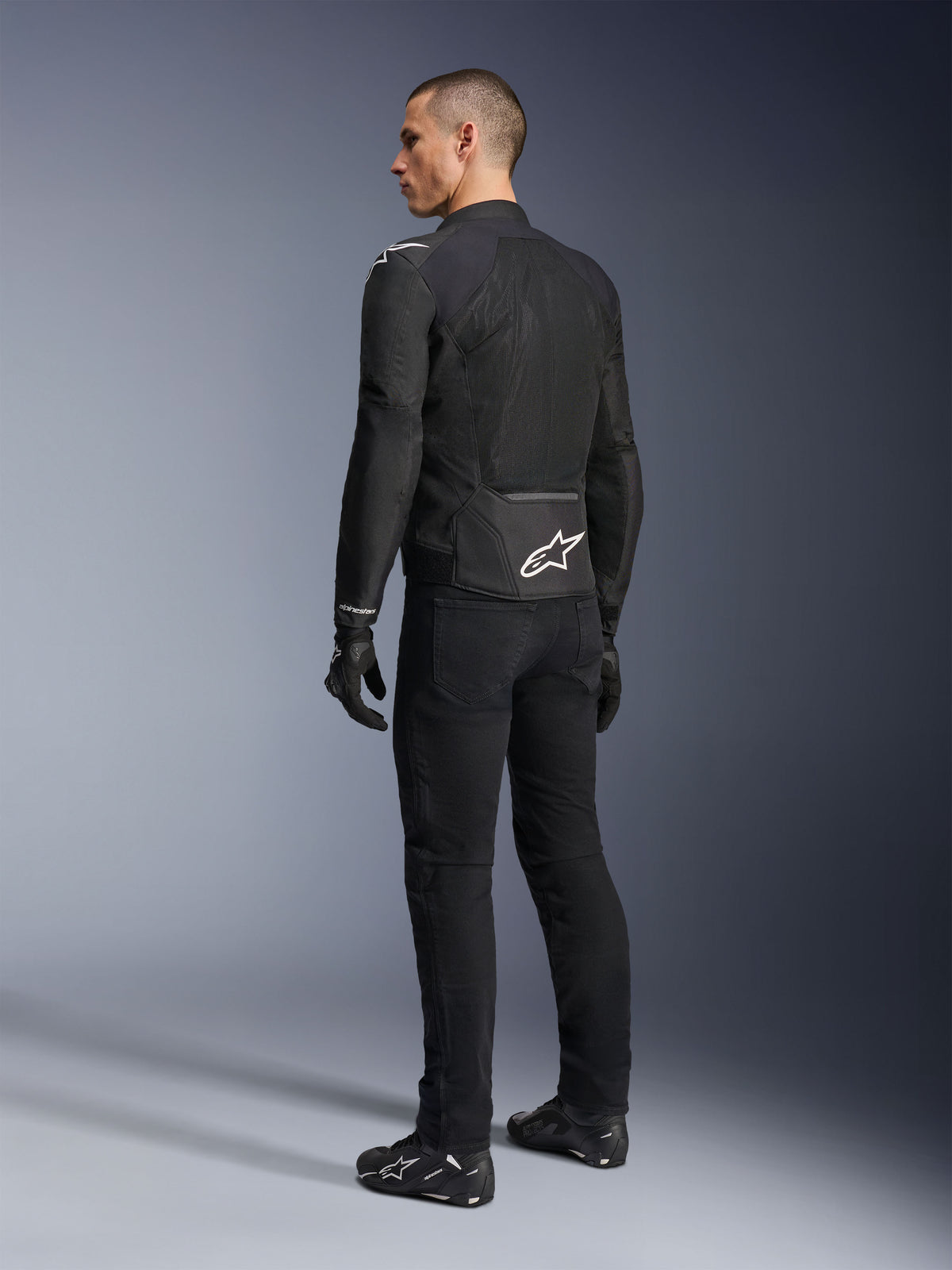 Kurtka Alpinestars Viper Air V4, czarna tekstylna kurtka motocyklowa, widok z tyłu na męskim modelu, duże panele z siatki technologicznej dla optymalnego przepływu powietrza, białe logo gwiazdy na dole pleców, noszona z czarnymi spodniami i rękawicami motocyklowymi
