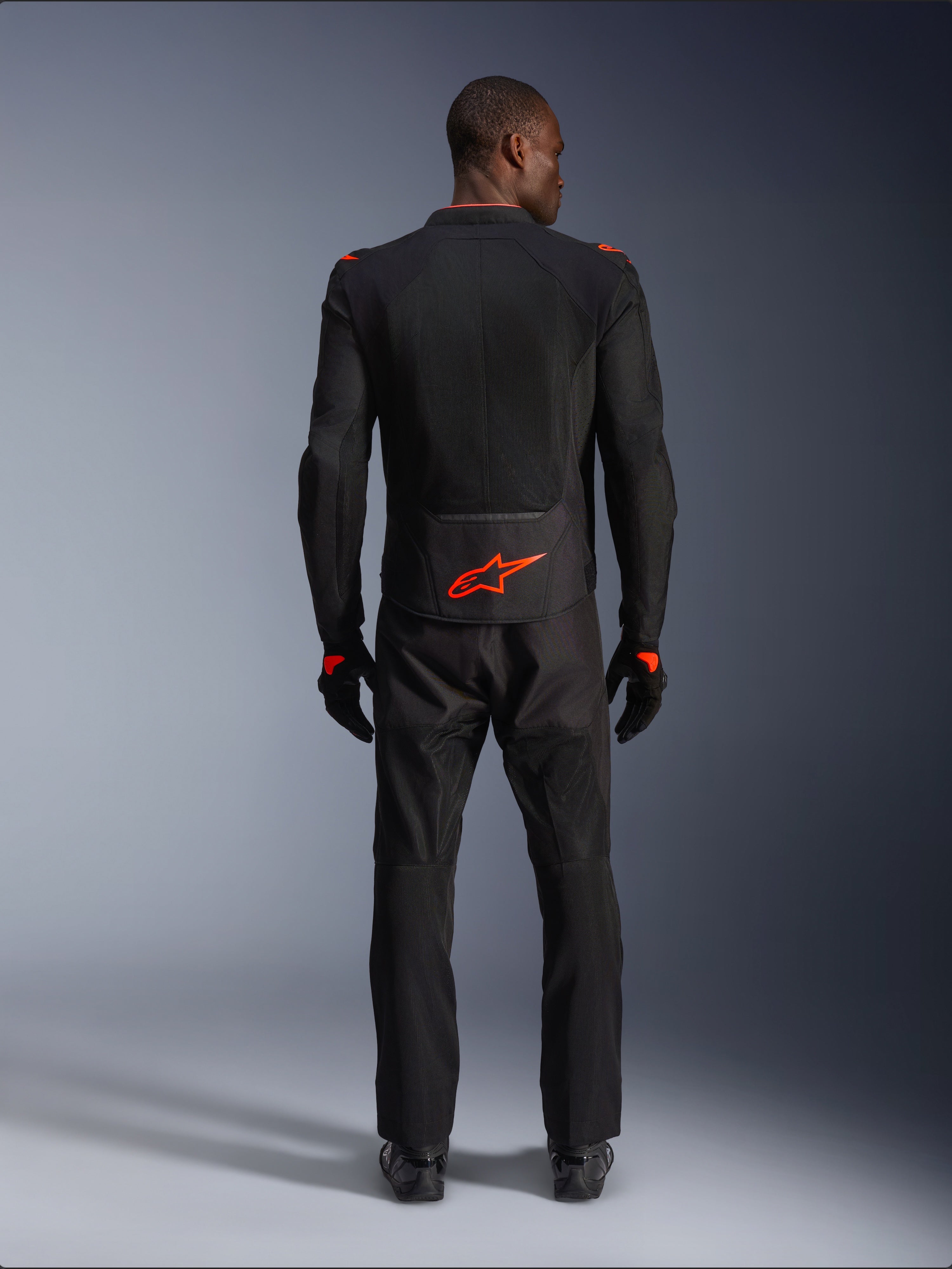 Kurtka Alpinestars Viper Air V4, Black Red Fluo, widok z tyłu na modelu, zestaw z dopasowanymi czarnymi spodniami i rękawicami motocyklowymi, jaskrawe neonowe czerwone logo na dole pleców, siateczkowa konstrukcja zapewniająca przepływ powietrza, sportowy krój do jazdy szosowej