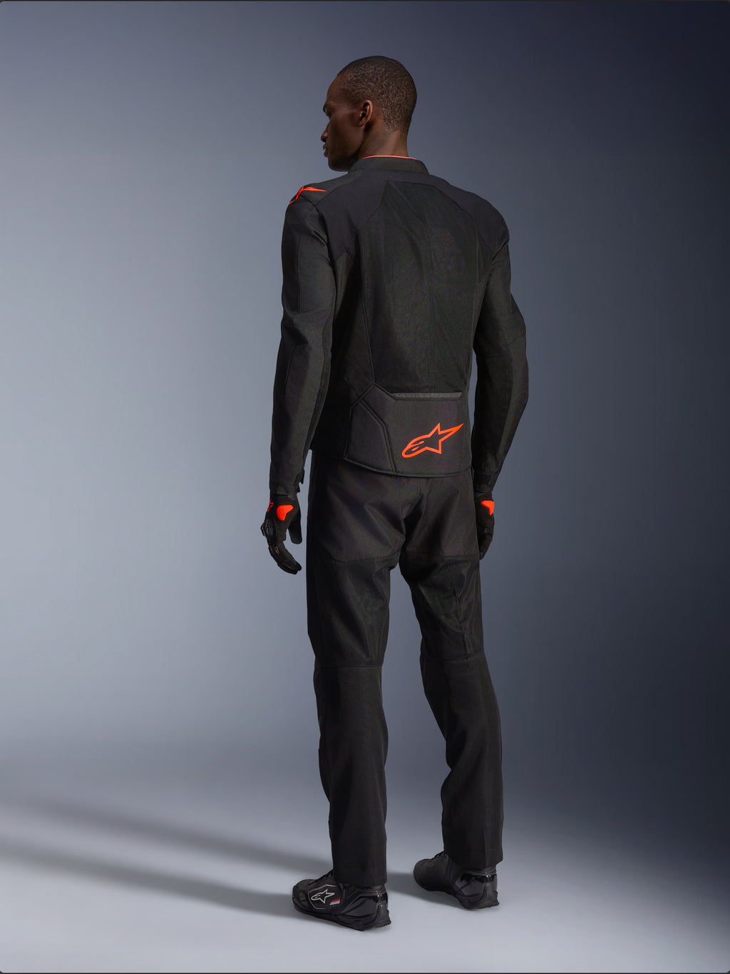 Alpinestars Viper Air V4 Jacket, motocyklista widziany od tyłu, czarno-czerwona (Black Red Fluo), pomarańczowe logo Astars na dole pleców i barkach, siatkowa konstrukcja tekstylna, czarne spodnie i buty motocyklowe