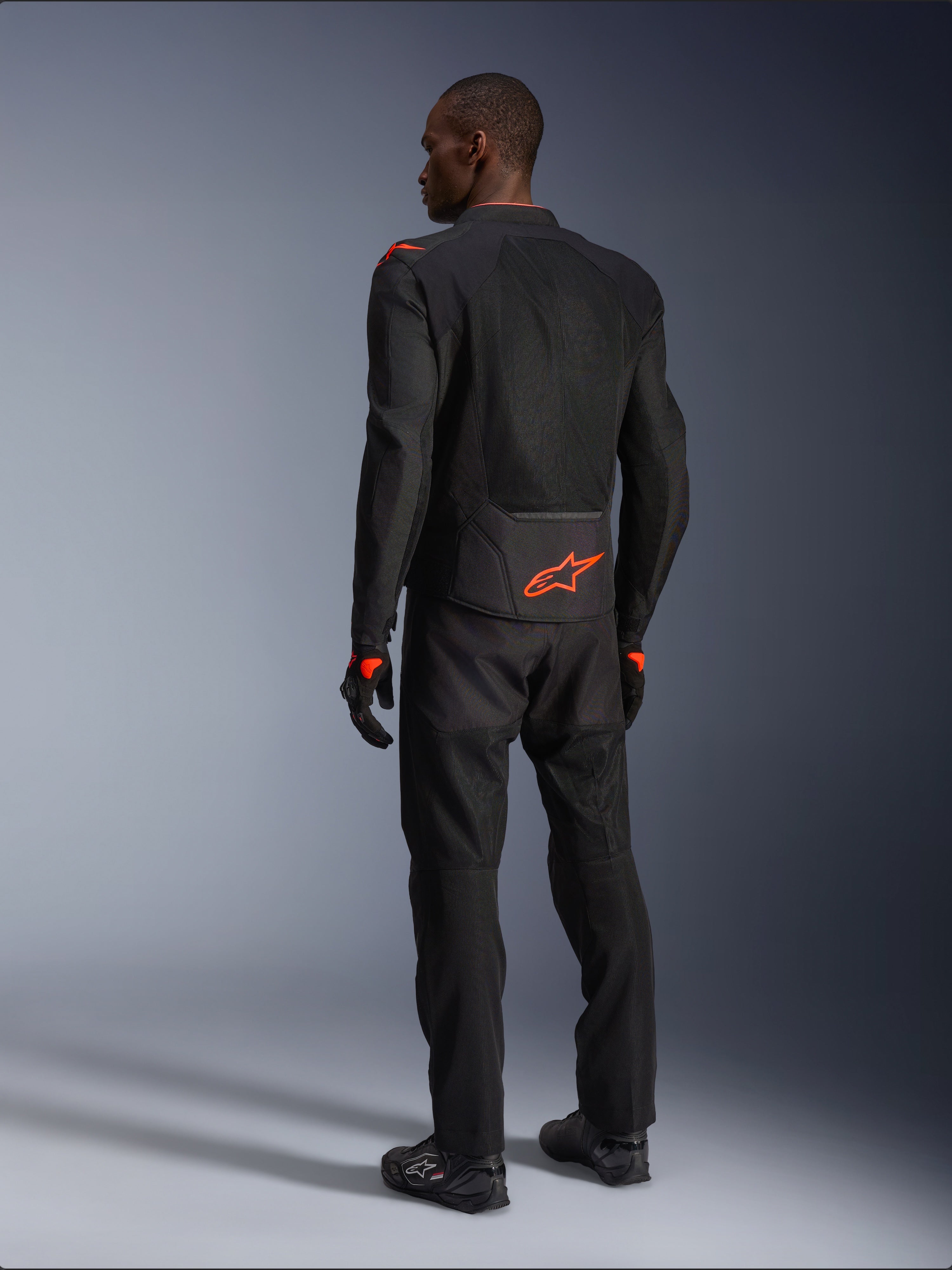Alpinestars Viper Air V4 Jacket, motocyklista widziany od tyłu, czarno-czerwona (Black Red Fluo), pomarańczowe logo Astars na dole pleców i barkach, siatkowa konstrukcja tekstylna, czarne spodnie i buty motocyklowe
