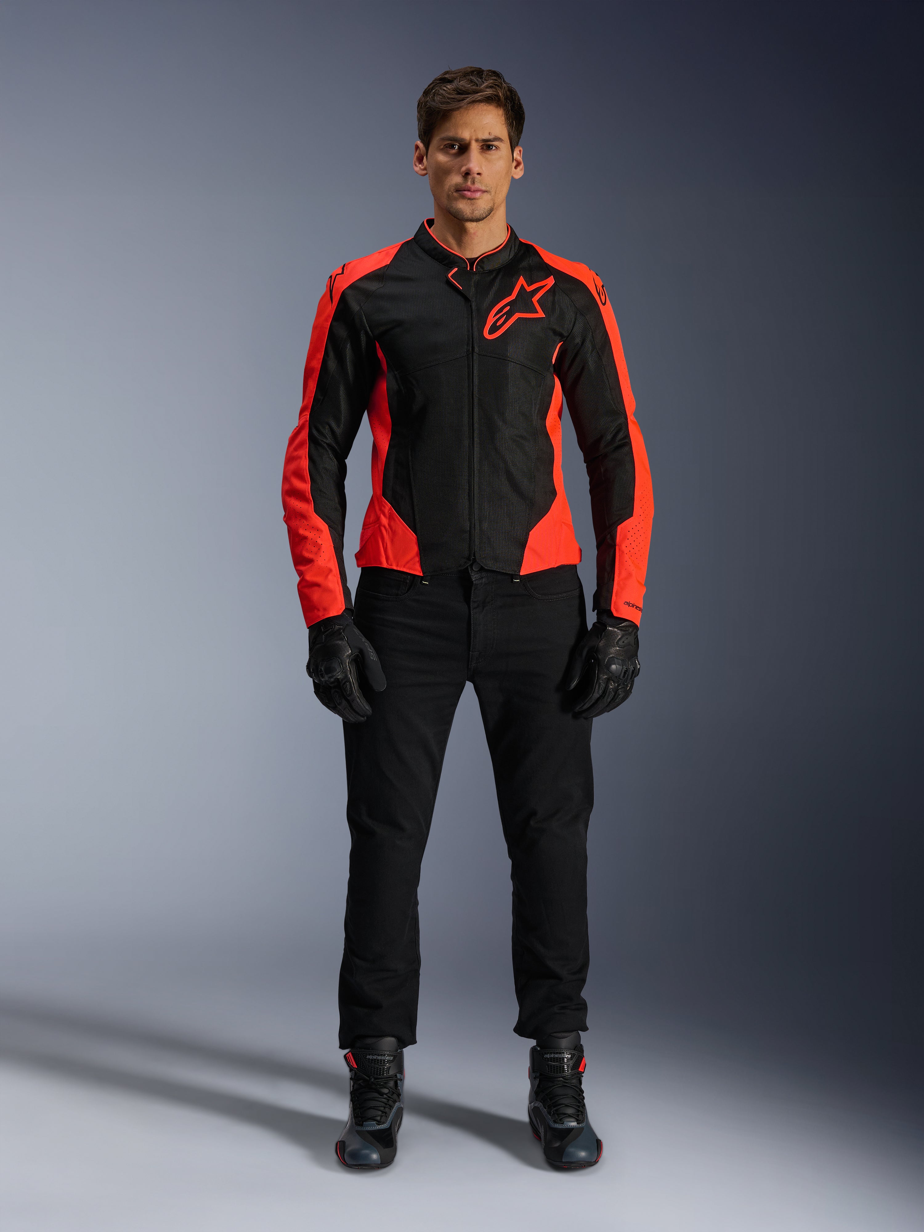 Męski model w kurtce Alpinestars Viper Air V4 w kolorze Red Fluo i czarnym, w zestawieniu z czarnymi spodniami, rękawicami i butami motocyklowymi, z oddychającymi panelami z siatki dla wentylacji i wyraźnym logo gwiazdy na piersi, sportowa odzież motocyklowa szosowa