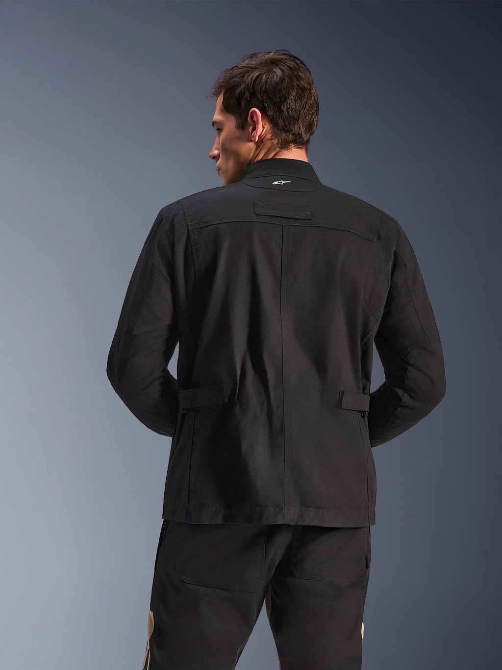 Alpinestars Dice Jacket, miejska kurtka motocyklowa, czarna, widok z tyłu prezentujący plisowane ramiona i regulowane patki w talii, estetyka kurtki polowej z małym białym logo na kołnierzu, ochronna odzież tekstylna do jazdy miejskiej