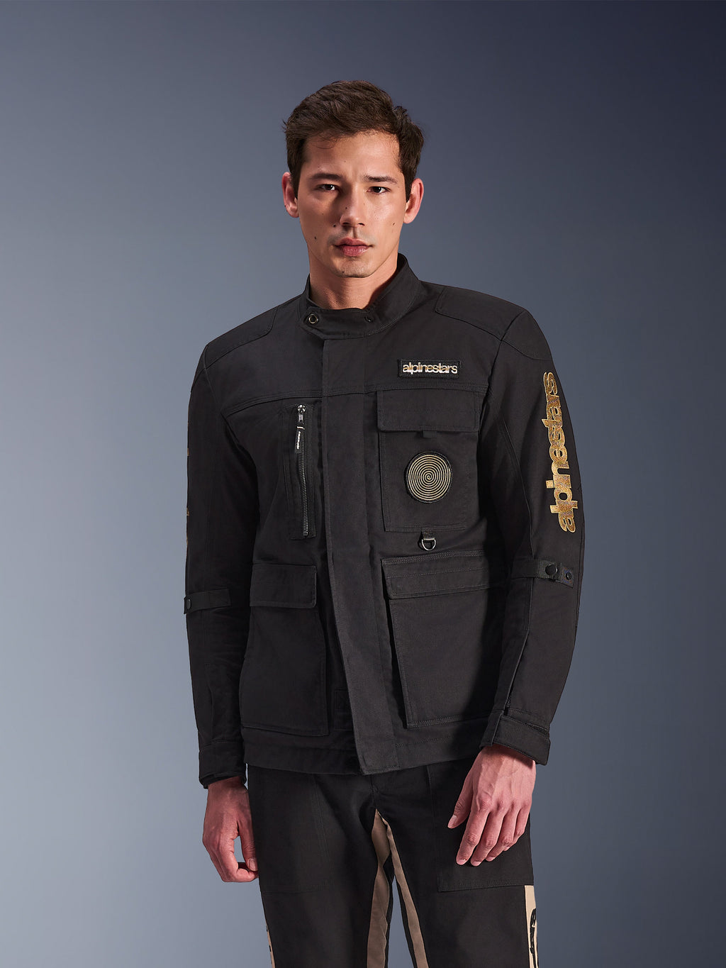 Alpinestars Dice Jacket, czarna kurtka motocyklowa, prezentowana przez modela, złote logo Alpinestars, liczne funkcjonalne kieszenie, okrągła naszywka ze spiralą, stylistyka miejskiej kurtki polowej