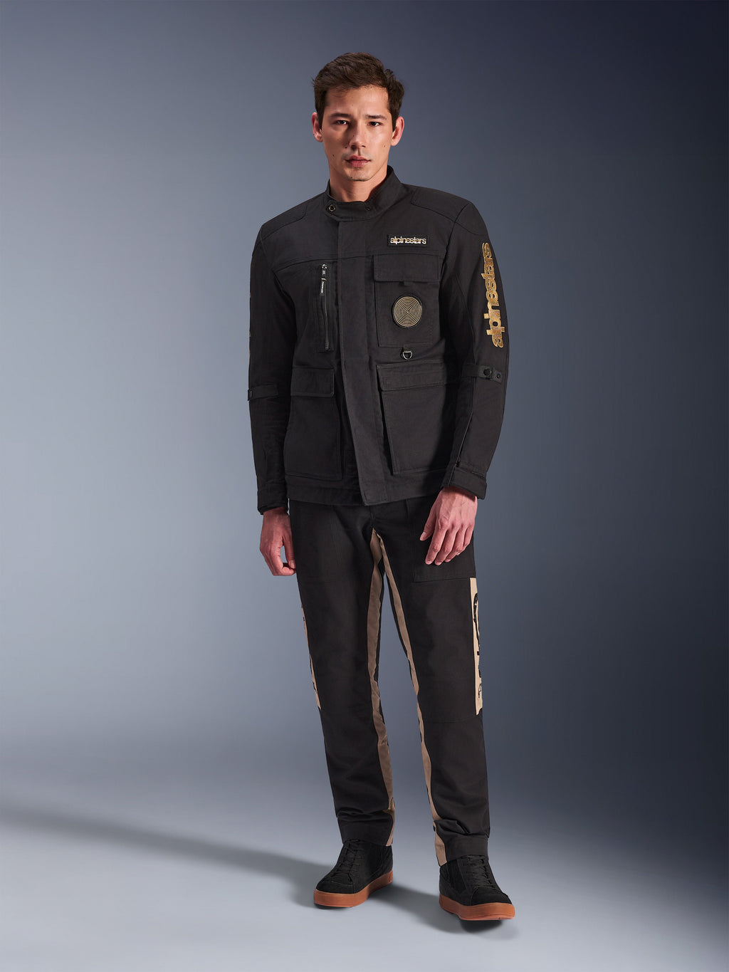 Męski model w kurtce Alpinestars Dice Jacket, czarna miejska kurtka motocyklowa w stylu vintage field jacket, cztery funkcjonalne kieszenie z przodu, branding Alpinestars na rękawie i piersi, zestawiona z czarno-beżowymi spodniami, nowoczesna miejska odzież motocyklowa