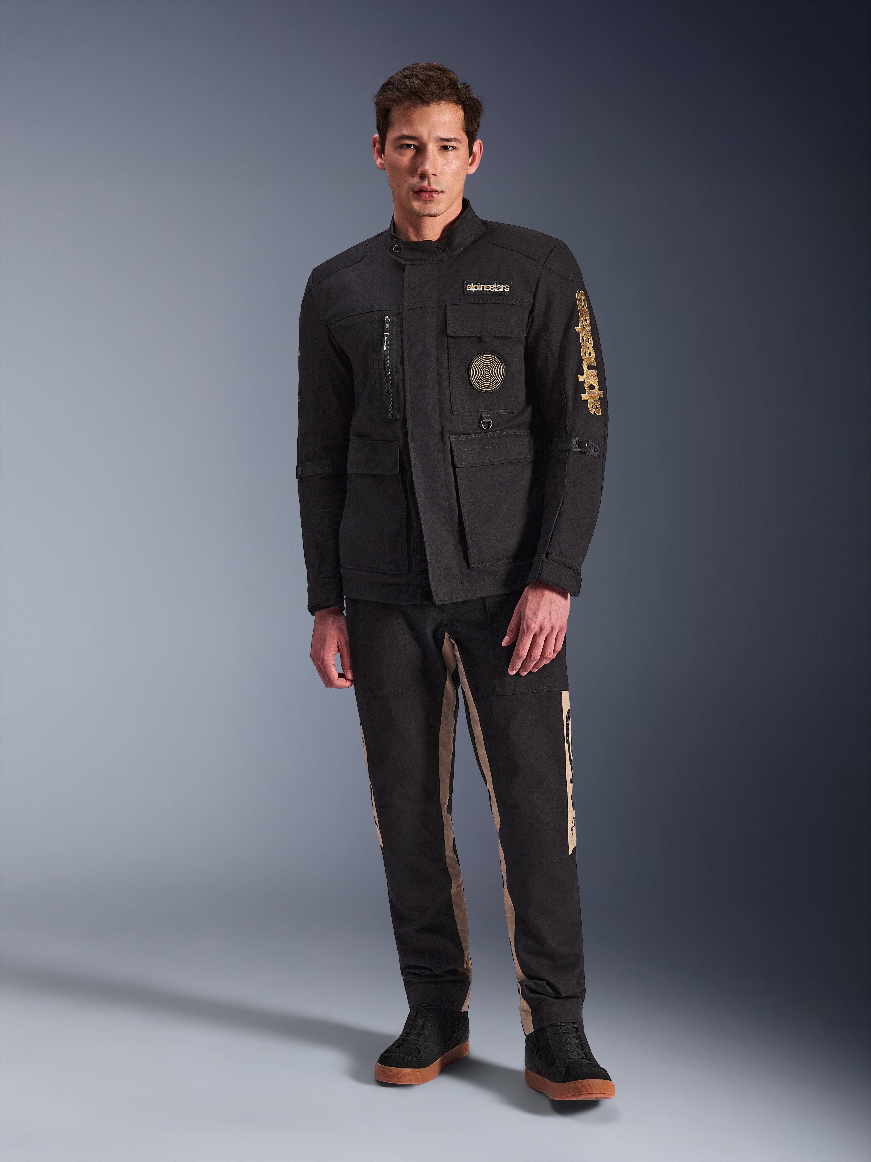Męski model w kurtce Alpinestars Dice Jacket, czarna miejska kurtka motocyklowa w stylu vintage field jacket, cztery funkcjonalne kieszenie z przodu, branding Alpinestars na rękawie i piersi, zestawiona z czarno-beżowymi spodniami, nowoczesna miejska odzież motocyklowa