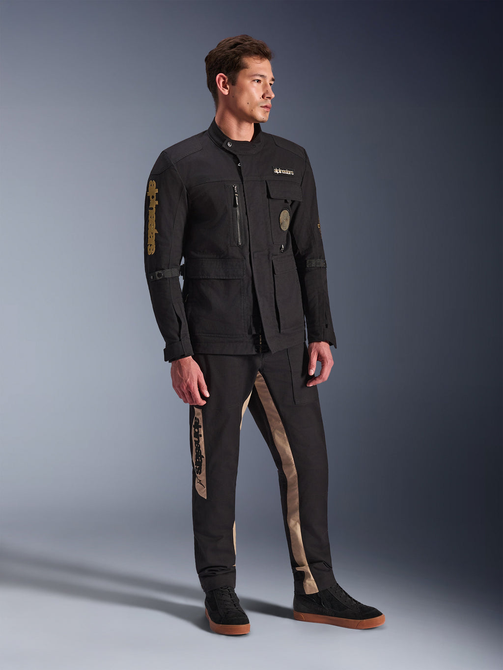 Męski model ubrany w czarną kurtkę Alpinestars Dice, miejską kurtkę motocyklową o estetyce vintage kurtki polowej, z licznymi kieszeniami i złotym brandingiem na rękawie, zestawioną z pasującymi czarno-beżowymi spodniami i czarnymi sneakersami.