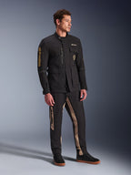 Męski model ubrany w czarną kurtkę Alpinestars Dice, miejską kurtkę motocyklową o estetyce vintage kurtki polowej, z licznymi kieszeniami i złotym brandingiem na rękawie, zestawioną z pasującymi czarno-beżowymi spodniami i czarnymi sneakersami.