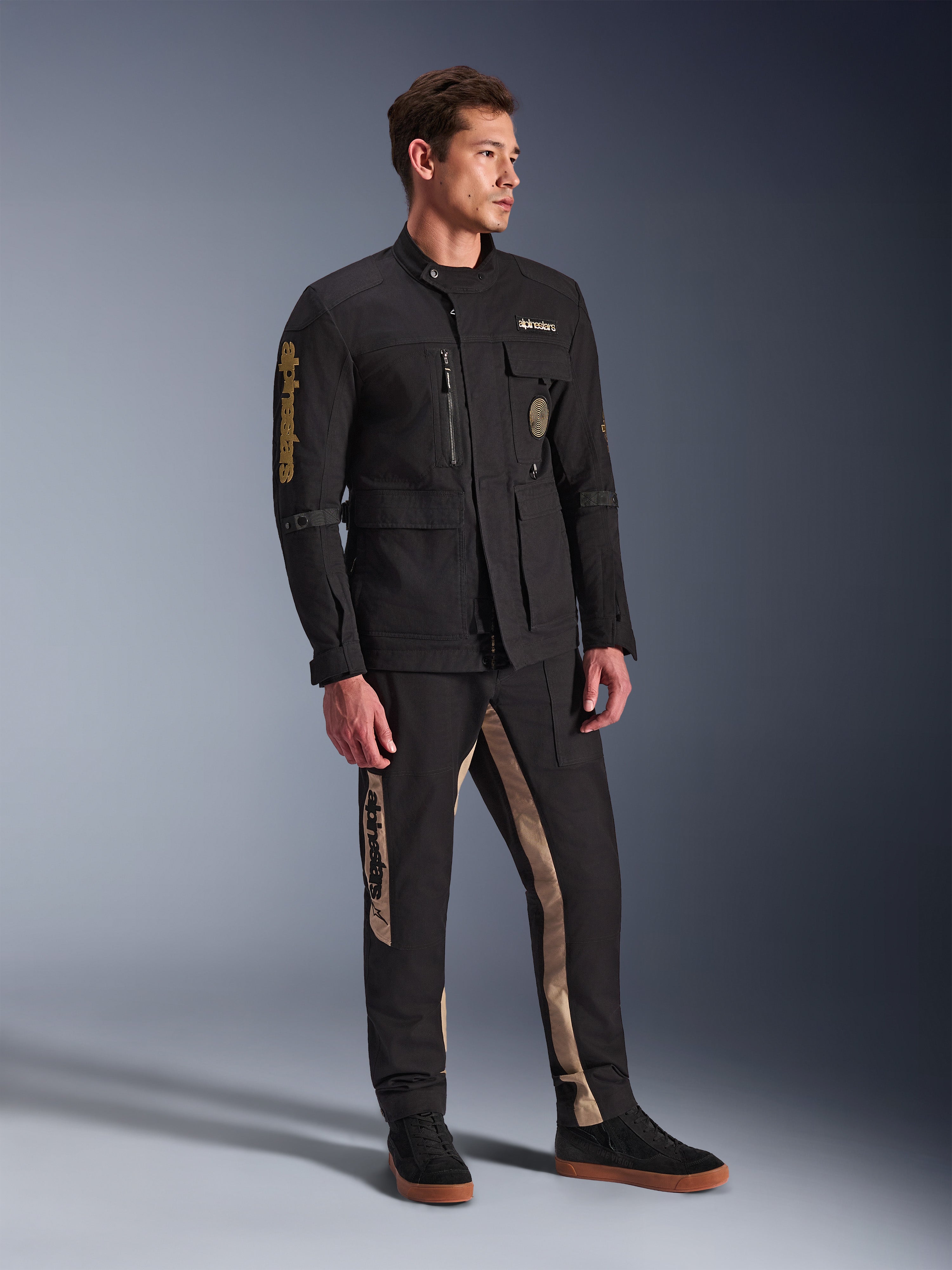 Męski model ubrany w czarną kurtkę Alpinestars Dice, miejską kurtkę motocyklową o estetyce vintage kurtki polowej, z licznymi kieszeniami i złotym brandingiem na rękawie, zestawioną z pasującymi czarno-beżowymi spodniami i czarnymi sneakersami.