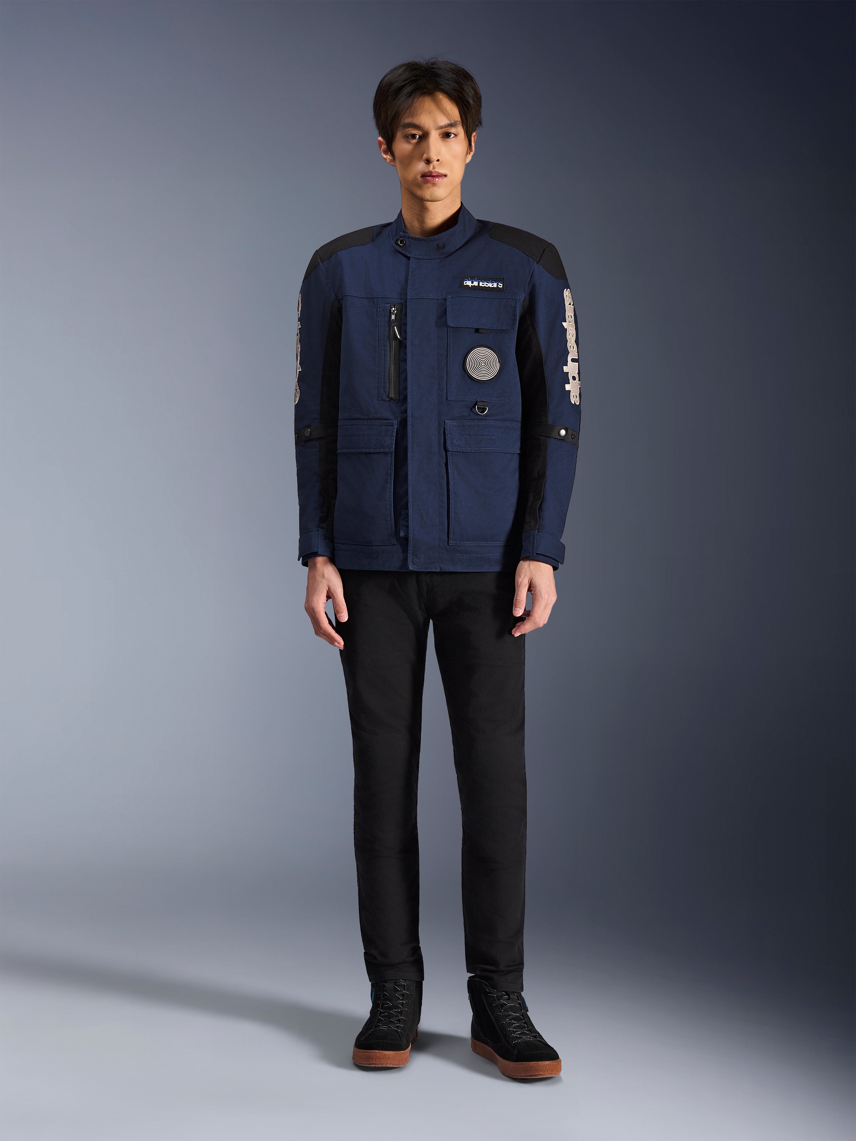 Alpinestars Dice Jacket, miejska kurtka motocyklowa, ciemnoniebieska i czarna, prezentowana na modelu z czarnymi spodniami i czarnymi butami, nowoczesny fason kurtki terenowej z wieloma kieszeniami, pionowy zamek na klatce piersiowej, białe logo Alpinestars na rękawach