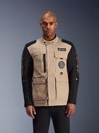 Alpinestars Dice Jacket, miejska kurtka motocyklowa, beżowo-czarna, surowa estetyka odzieży roboczej z licznymi kieszeniami użytkowymi i naszywkami na piersi, wysoki kołnierz zapinany na guzik, wyraziste logo na rękawach, prezentowana przez modela