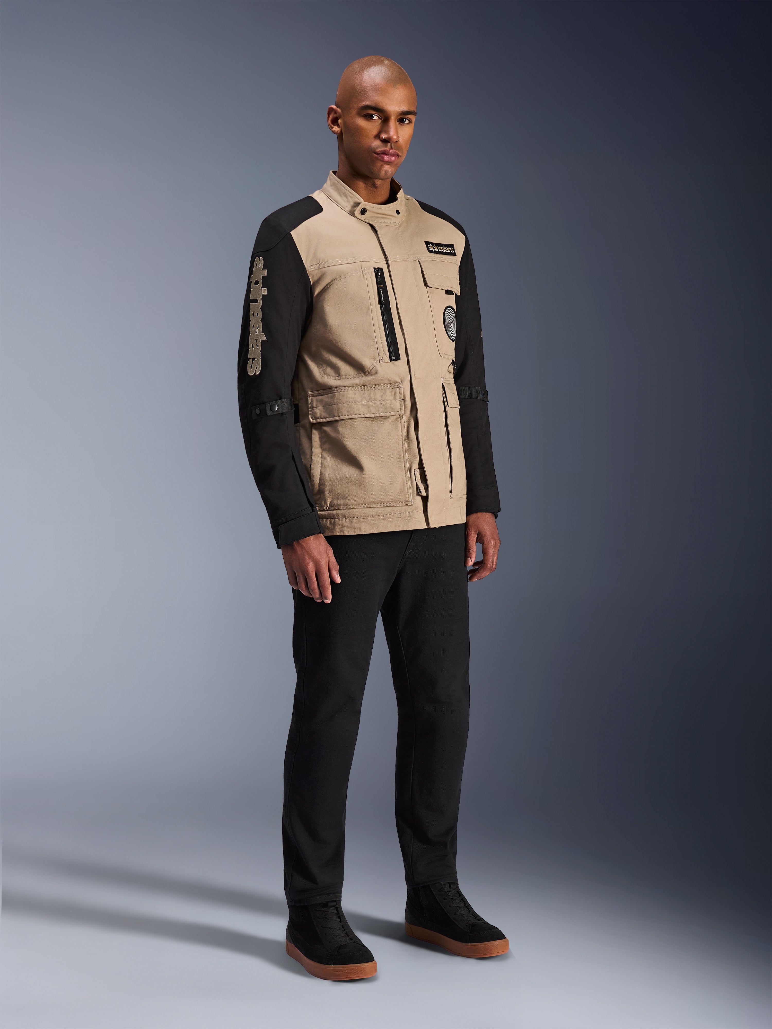 Alpinestars Dice Jacket, miejska kurtka motocyklowa, czarno-piaskowa, dwukolorowy design z czarnymi rękawami i korpusem w kolorze khaki, noszona przez modela w czarnych spodniach i czarnych butach sportowych, estetyka inspirowana odzieżą roboczą z kieszeniami użytkowymi i haftowanym logo na rękawie.