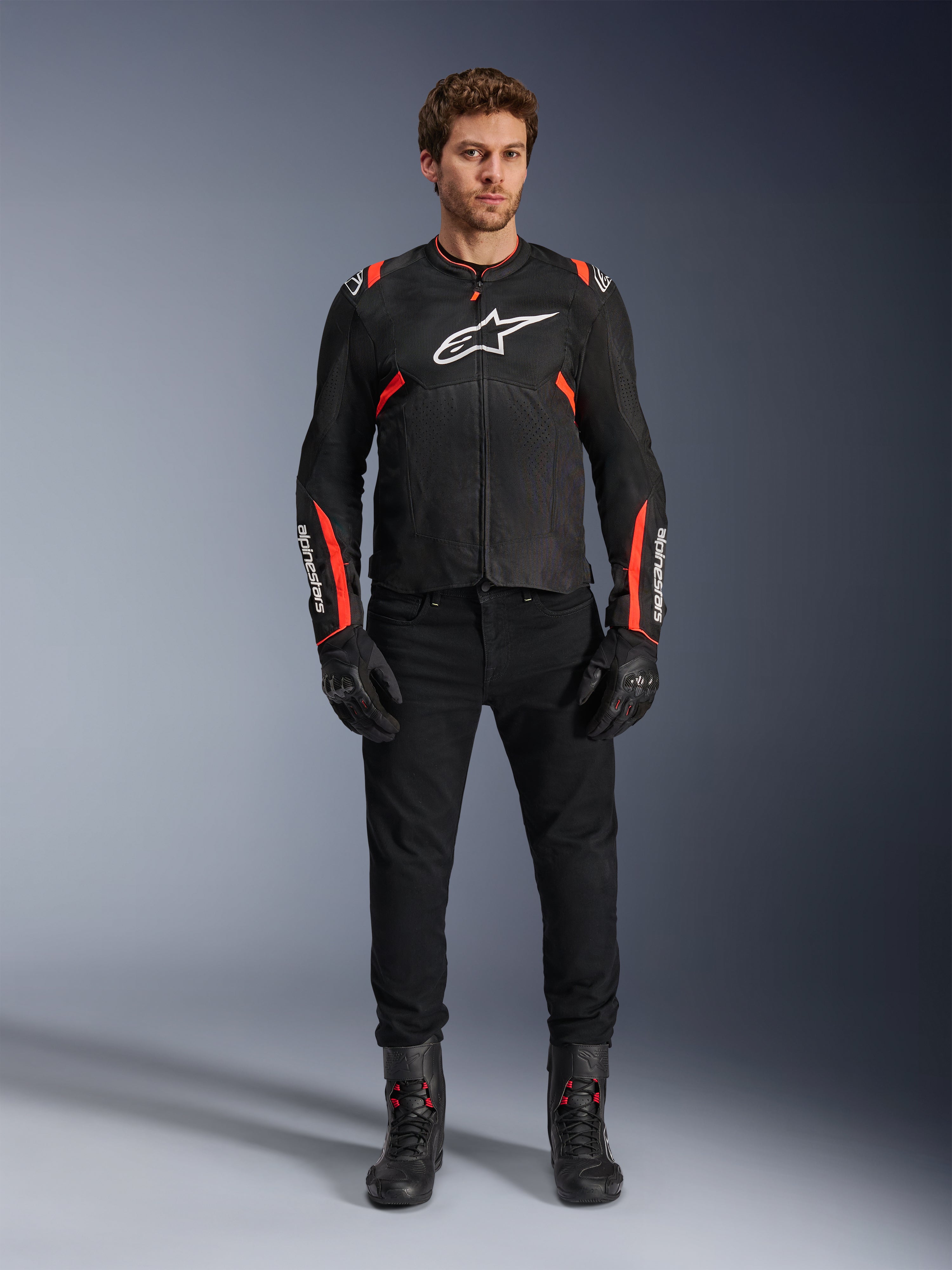 Męski model w kurtce Alpinestars T-SPS Air v2, czarna tekstylna kurtka motocyklowa z akcentami Red Fluo, białe logo na piersi, noszona z czarnymi spodniami, rękawicami i butami, miejski sportowy styl jazdy