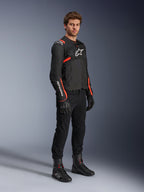 Męski model ubrany w kurtkę Alpinestars T-Sps Air V2, czarną z akcentami Red Fluo, w zestawieniu z czarnymi spodniami motocyklowymi, czarnymi rękawicami i czarnymi butami, podkreślającą sportowy design z panelami z siatki i białym logo Alpinestars.