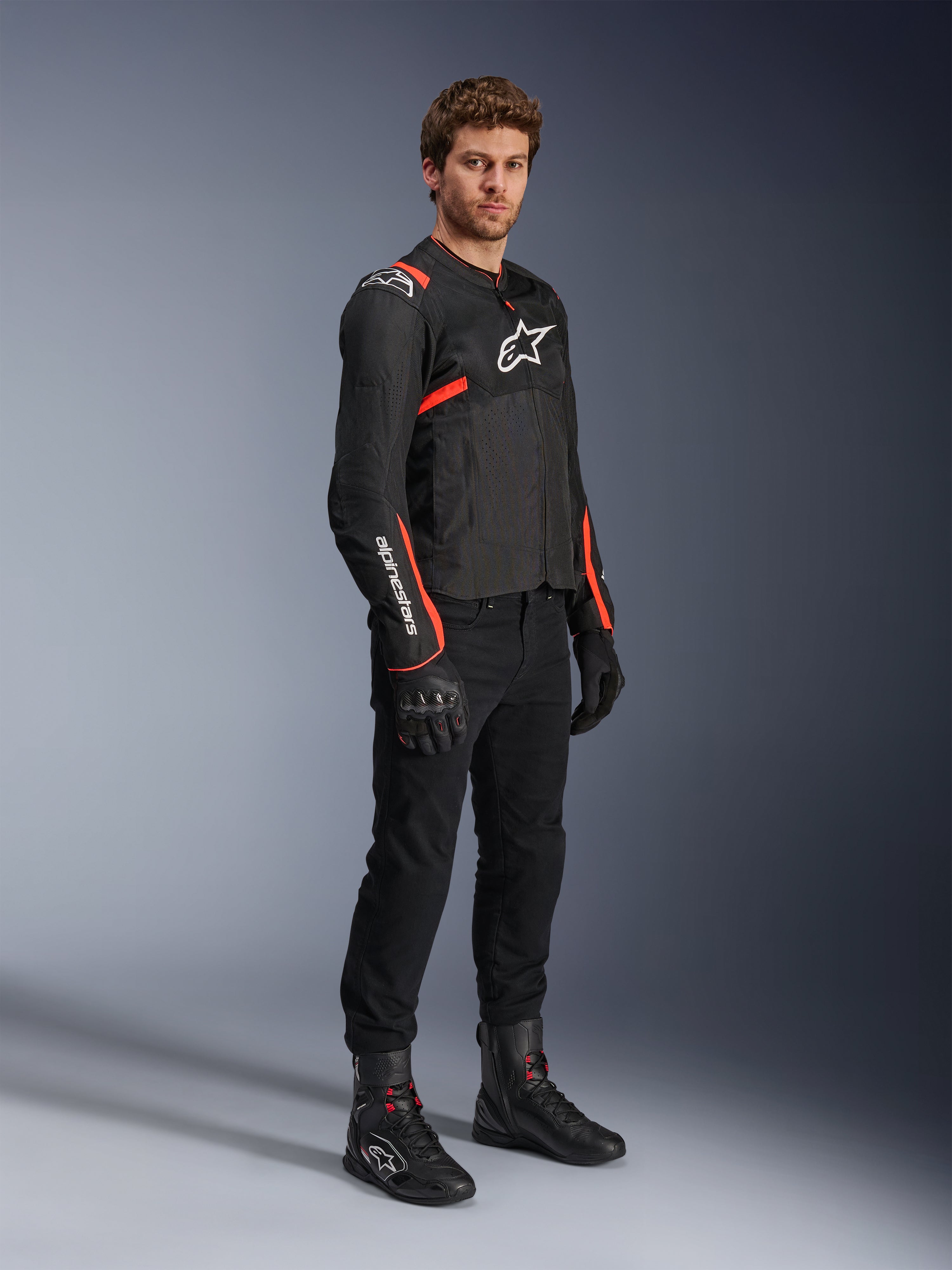 Męski model ubrany w kurtkę Alpinestars T-Sps Air V2, czarną z akcentami Red Fluo, w zestawieniu z czarnymi spodniami motocyklowymi, czarnymi rękawicami i czarnymi butami, podkreślającą sportowy design z panelami z siatki i białym logo Alpinestars.