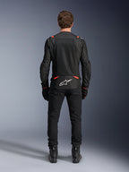 Kurtka Alpinestars T-SPS Air v2 w kolorze czarnym i czerwonym fluo, widok z tyłu przedstawiający duże siatkowe panele wentylacyjne i białe logo Alpinestars na dole pleców, zestawiona z czarnymi spodniami i butami motocyklowymi.