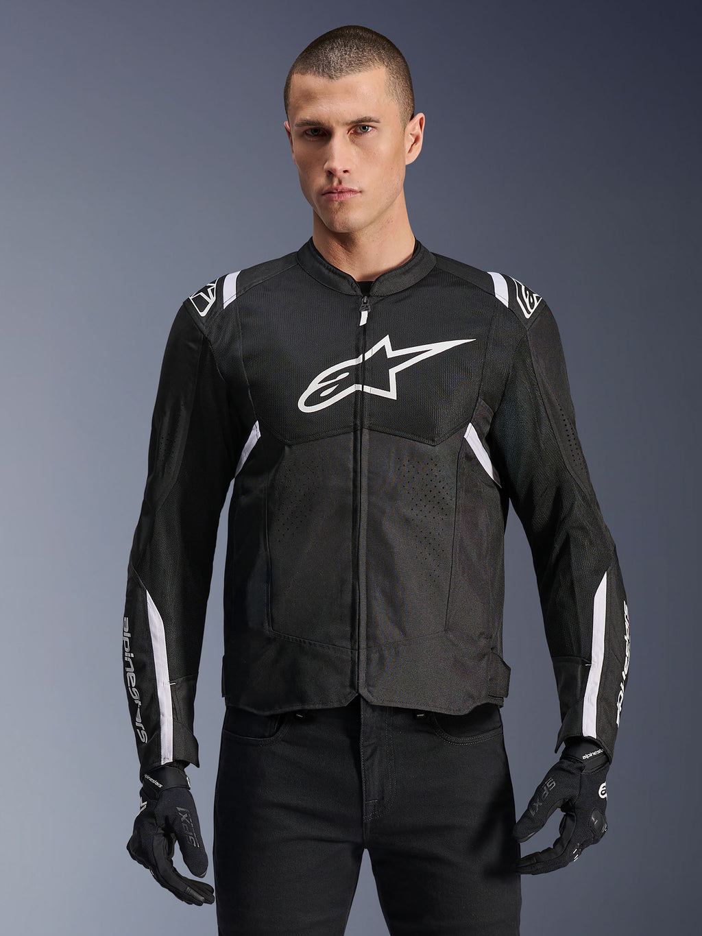 Alpinestars T-SPS Air V2 Jacket, tekstylna kurtka motocyklowa, czarna z białymi akcentami, duże białe logo na piersi, perforowana konstrukcja z siatki dla wentylacji, noszona z czarnymi spodniami i rękawicami