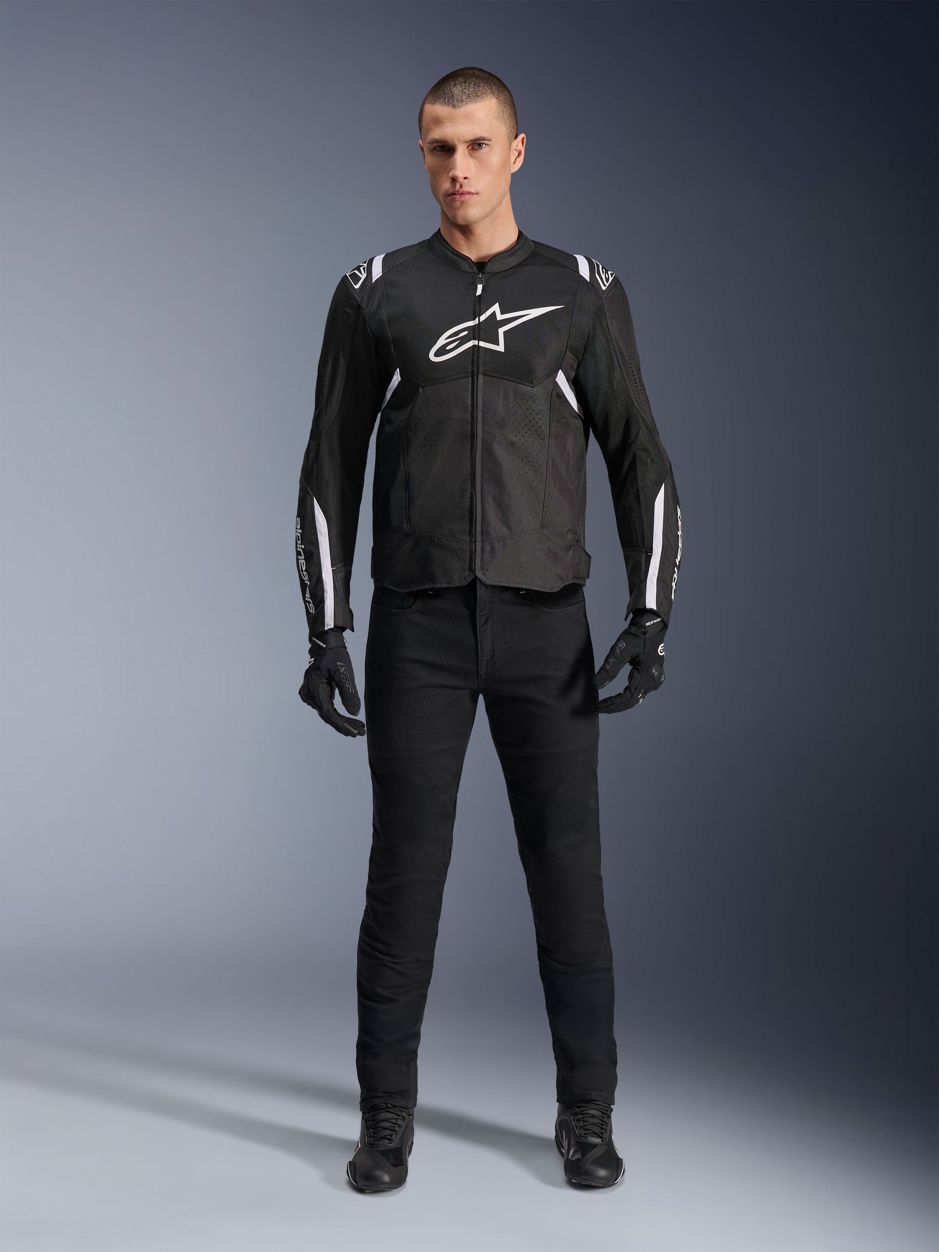 Alpinestars T-Sps Air V2 Jacket, czarno-biała tekstylna kurtka motocyklowa z dużym białym logo na klatce piersiowej i panelami z siatki, noszona przez modela w czarnym stroju do sportowej jazdy miejskiej
