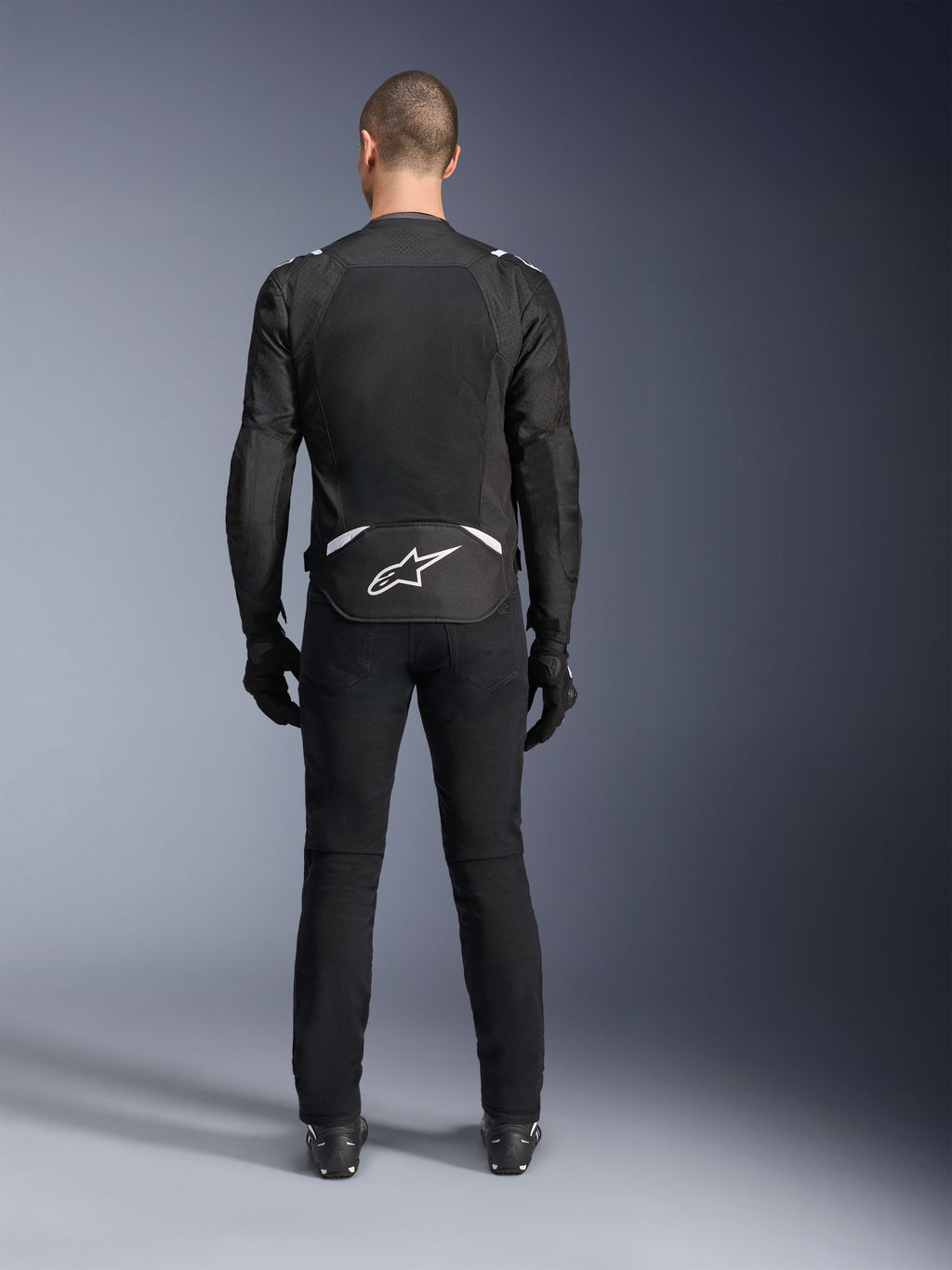 Alpinestars T-Sps Air V2 Jacket, widok z tyłu, czarno-biała tekstylna kurtka motocyklowa, duży panel z siatki na plecach zapewniający maksymalny przepływ powietrza, białe logo Astars w dolnej części pleców, zestawiona z czarnymi spodniami i rękawicami