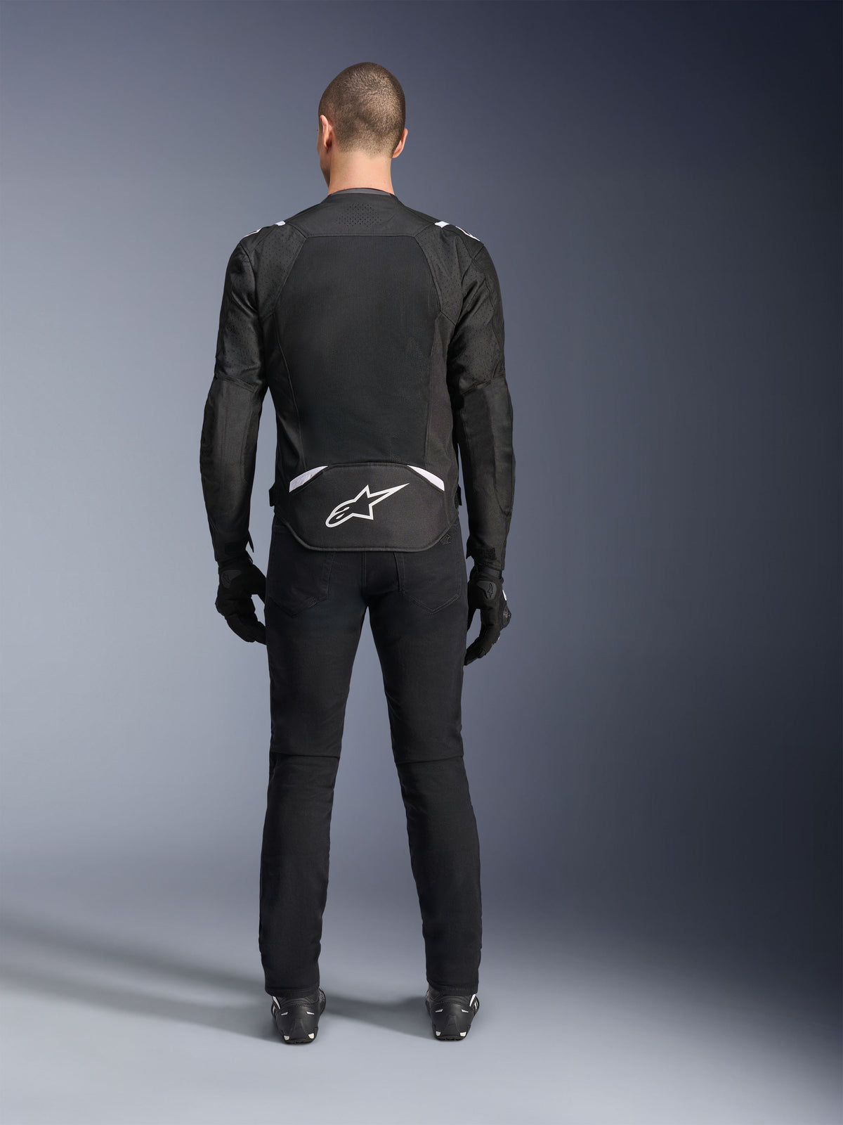 Alpinestars T-Sps Air V2 Jacket, widok z tyłu, czarno-biała tekstylna kurtka motocyklowa, duży panel z siatki na plecach zapewniający maksymalny przepływ powietrza, białe logo Astars w dolnej części pleców, zestawiona z czarnymi spodniami i rękawicami