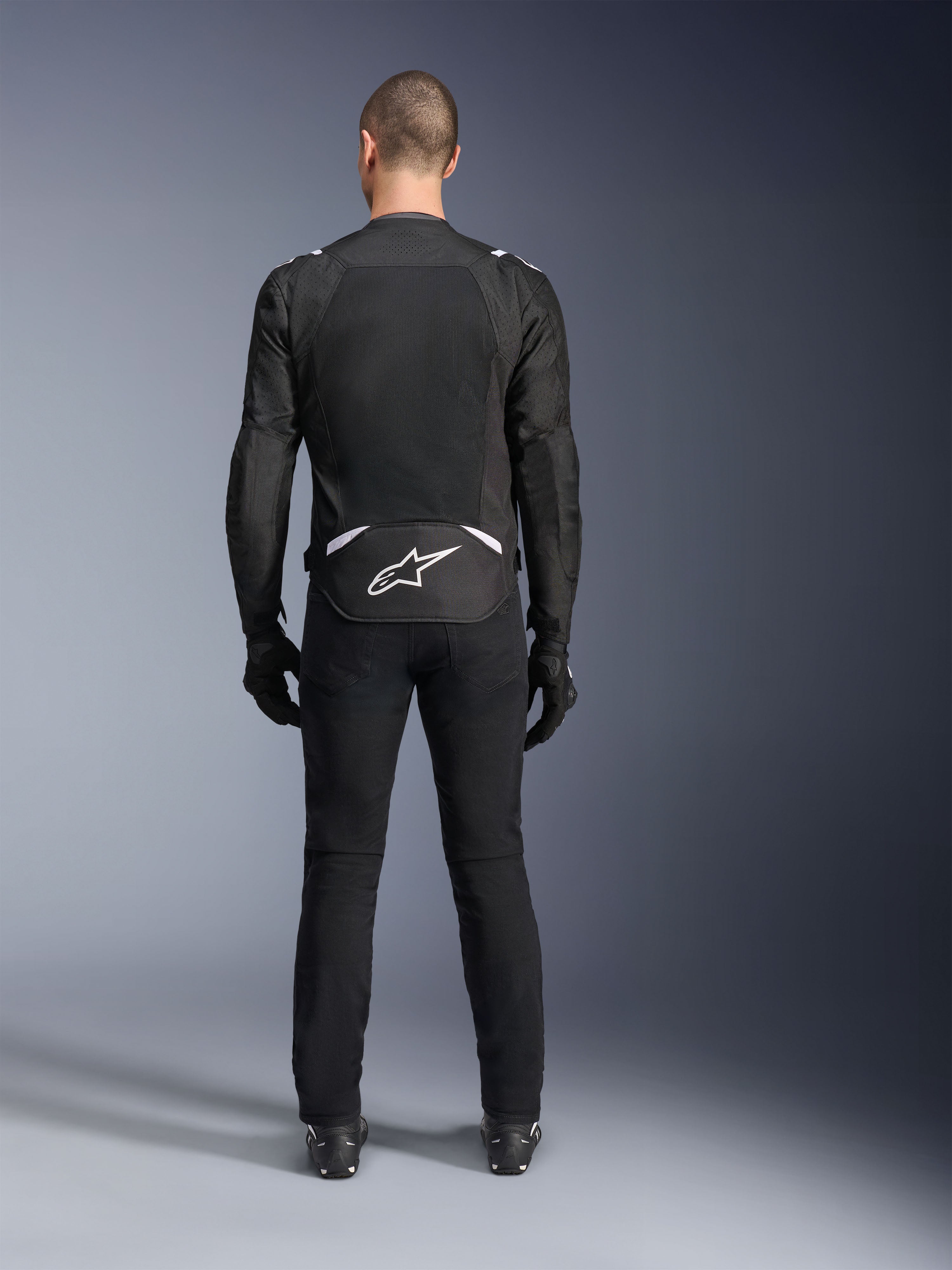 Alpinestars T-Sps Air V2 Jacket, widok z tyłu, czarno-biała tekstylna kurtka motocyklowa, duży panel z siatki na plecach zapewniający maksymalny przepływ powietrza, białe logo Astars w dolnej części pleców, zestawiona z czarnymi spodniami i rękawicami