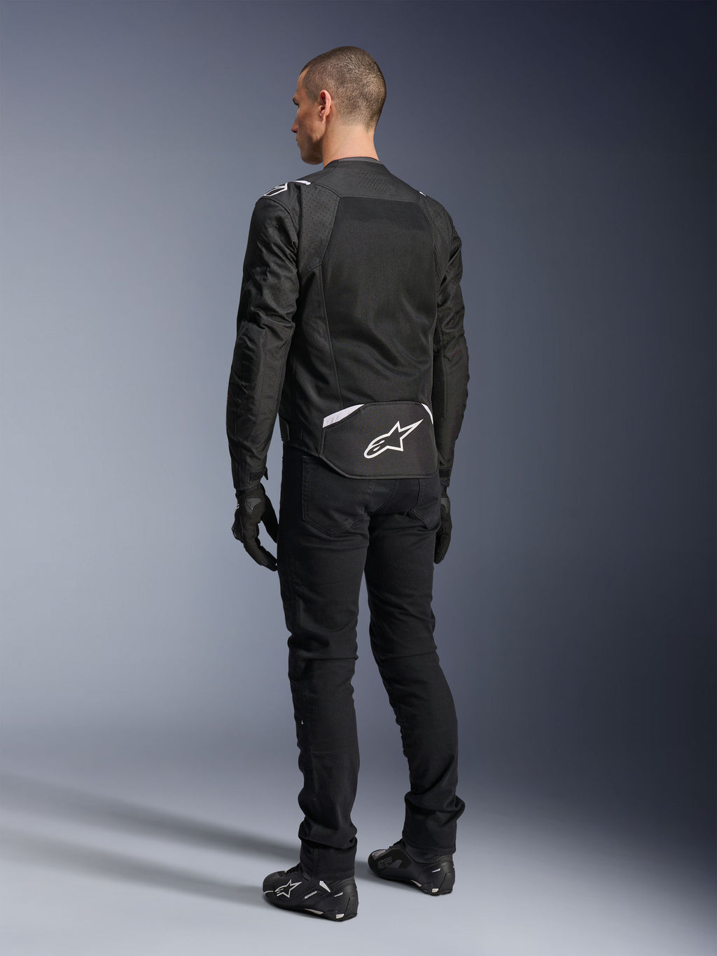 Kurtka Alpinestars T-Sps Air V2, tekstylna kurtka motocyklowa, czarna, widok z tyłu ukazujący duży panel wentylacyjny z siatki oraz białe logo Alpinestars w dolnej części pleców, zestawiona z czarnymi spodniami i rękawicami motocyklowymi dla sportowego, miejskiego stylu.