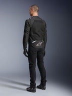Kurtka Alpinestars T-Sps Air V2, tekstylna kurtka motocyklowa, czarna, widok z tyłu ukazujący duży panel wentylacyjny z siatki oraz białe logo Alpinestars w dolnej części pleców, zestawiona z czarnymi spodniami i rękawicami motocyklowymi dla sportowego, miejskiego stylu.