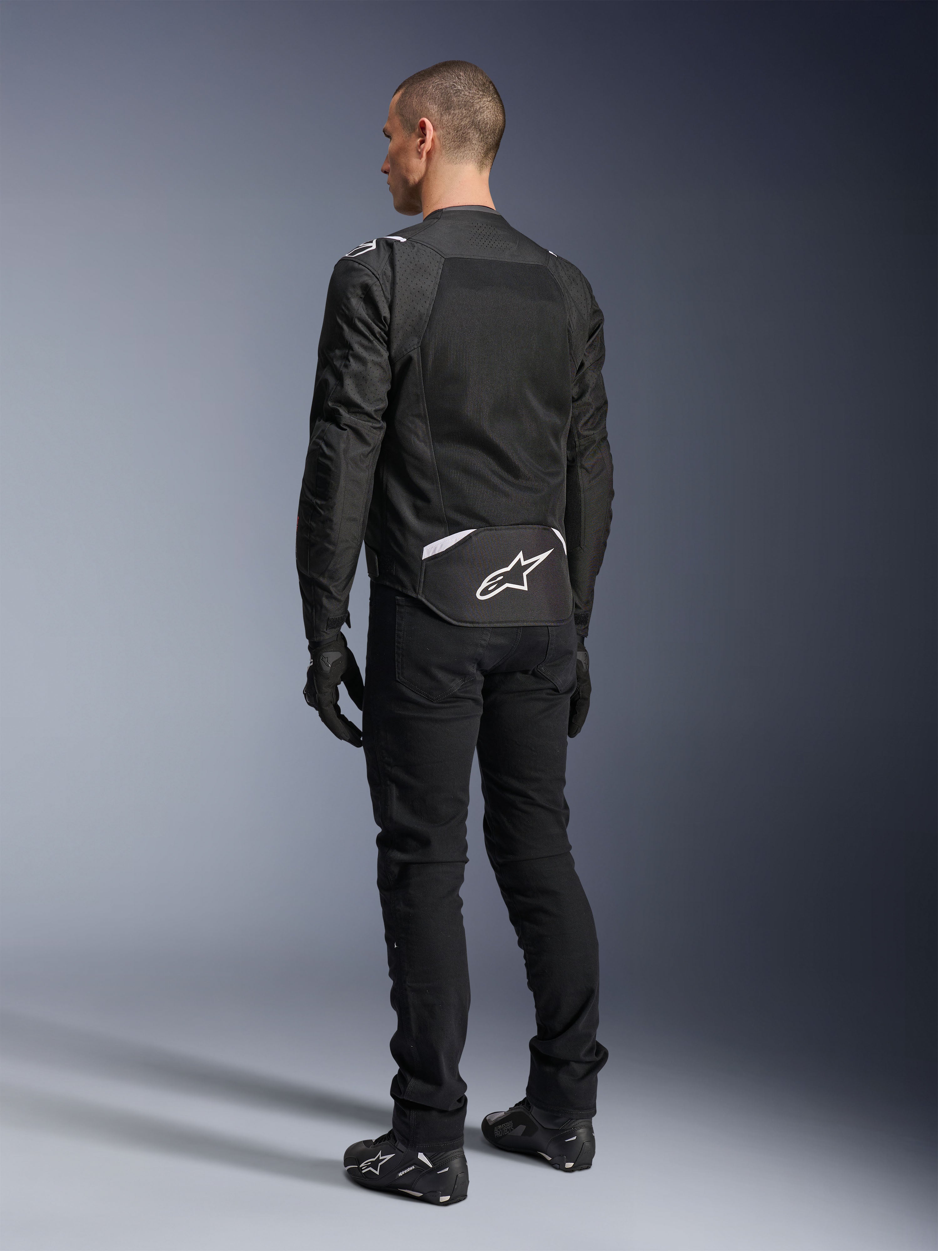 Kurtka Alpinestars T-Sps Air V2, tekstylna kurtka motocyklowa, czarna, widok z tyłu ukazujący duży panel wentylacyjny z siatki oraz białe logo Alpinestars w dolnej części pleców, zestawiona z czarnymi spodniami i rękawicami motocyklowymi dla sportowego, miejskiego stylu.