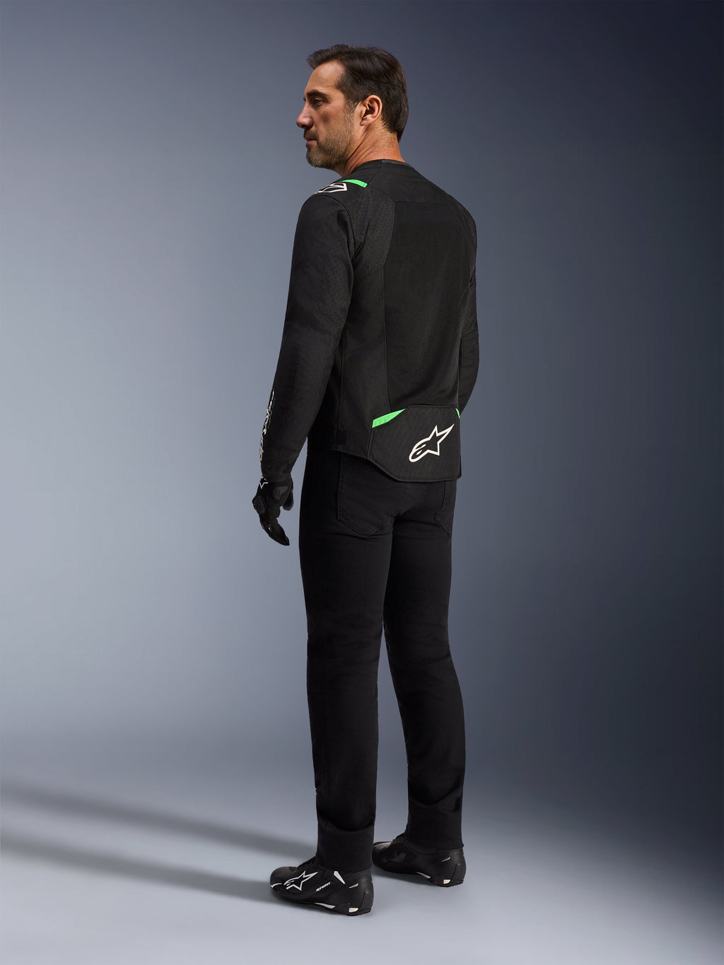 Mężczyzna w kurtce Alpinestars T-SPS Air V2 w kolorze Black Fluo Green, widoczny z tyłu i z profilu, w czarnych spodniach do jazdy i butach motocyklowych. Kurtka posiada wentylowane panele z siatki technicznej, jaskrawozielone akcenty na ramionach i w dolnej części pleców oraz białe logo gwiazdy na dole pleców.