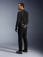 Mężczyzna w kurtce Alpinestars T-SPS Air V2 w kolorze Black Fluo Green, widoczny z tyłu i z profilu, w czarnych spodniach do jazdy i butach motocyklowych. Kurtka posiada wentylowane panele z siatki technicznej, jaskrawozielone akcenty na ramionach i w dolnej części pleców oraz białe logo gwiazdy na dole pleców.