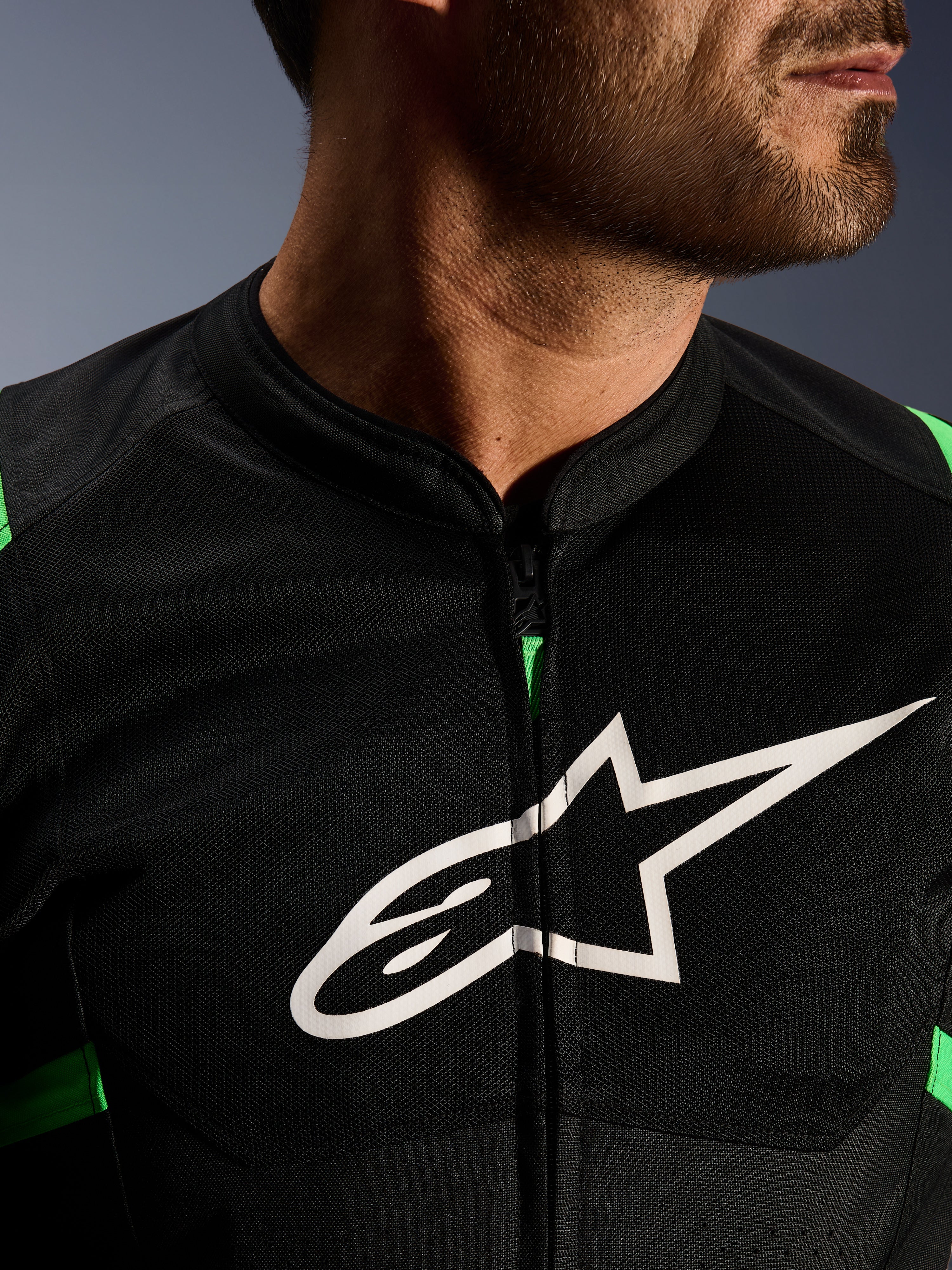 Zbliżenie na kurtkę Alpinestars T-SPS Air V2, czarno-zielona (Fluo Green), z oddychającymi panelami tech mesh, centralnym zamkiem i wyraźnym białym logo gwiazdy Alpinestars na klatce piersiowej, wyczynowa odzież motocyklowa