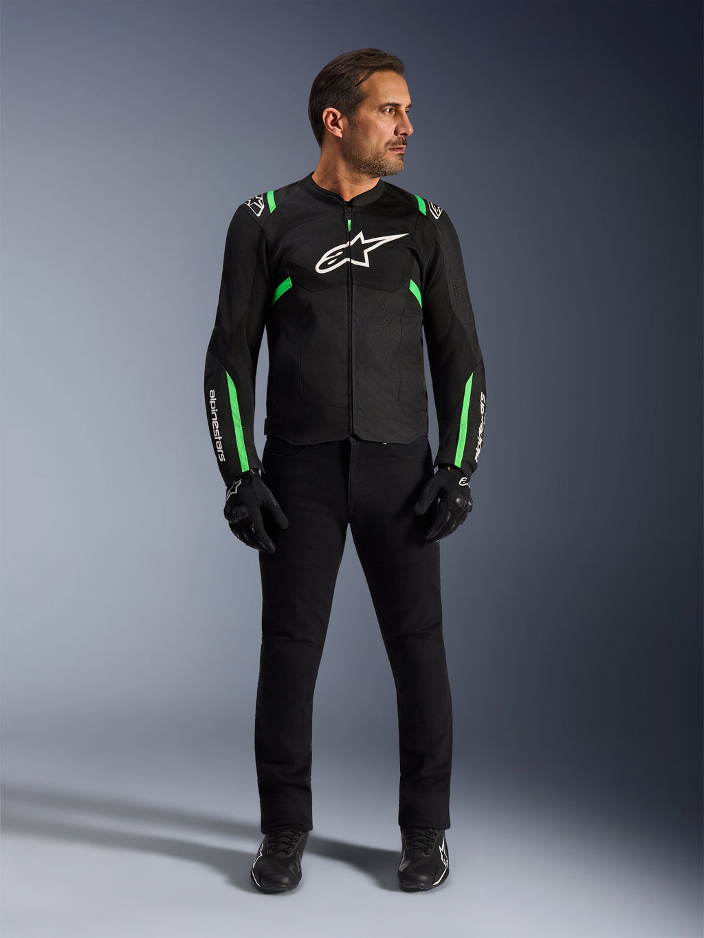 Mężczyzna noszący kurtkę Alpinestars T-SPS Air V2 w kolorze Black Fluo Green, zestawioną z czarnymi spodniami, rękawicami i butami motocyklowymi, o sportowym kroju z białymi logo i panelami z siatki, zapewniający nowoczesny miejski wygląd motocyklowy.