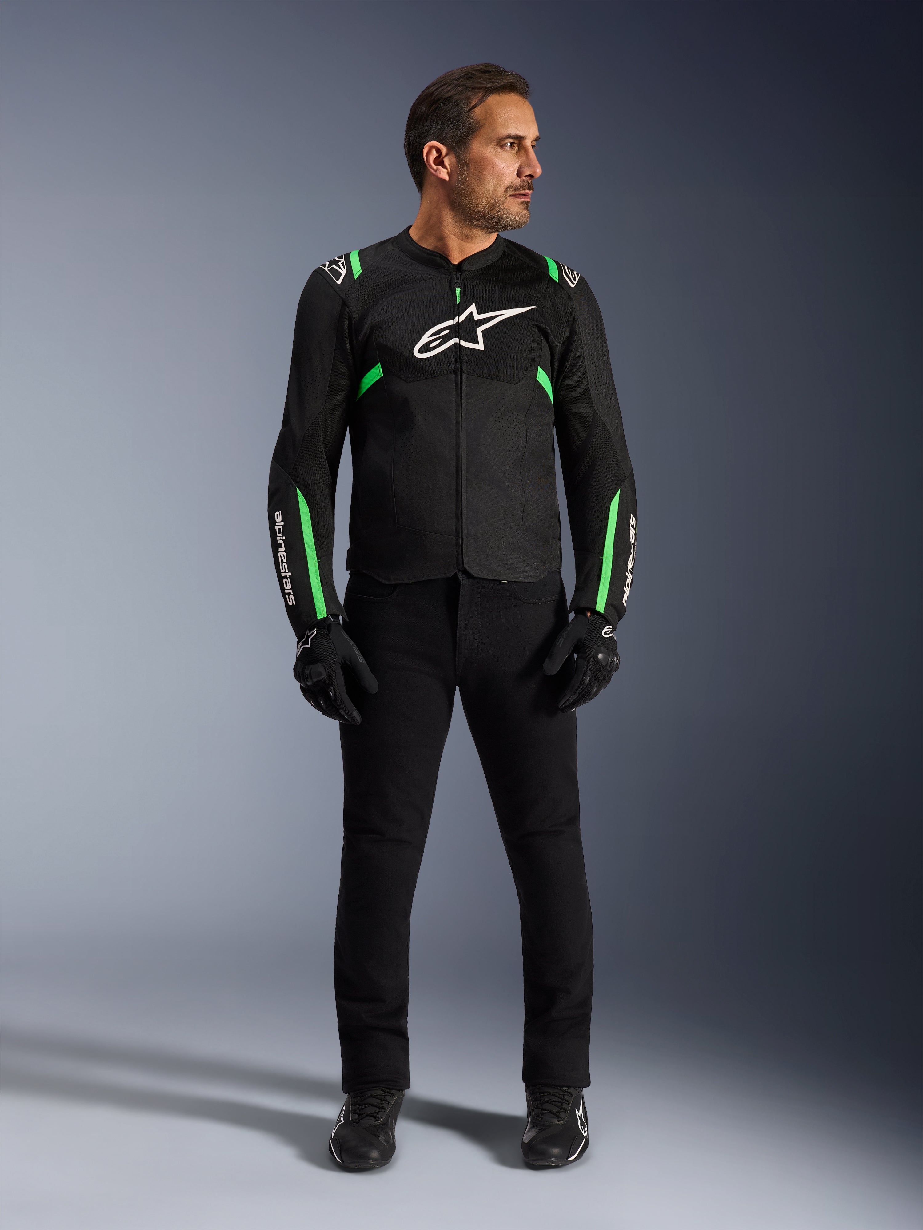 Mężczyzna noszący kurtkę Alpinestars T-SPS Air V2 w kolorze Black Fluo Green, zestawioną z czarnymi spodniami, rękawicami i butami motocyklowymi, o sportowym kroju z białymi logo i panelami z siatki, zapewniający nowoczesny miejski wygląd motocyklowy.